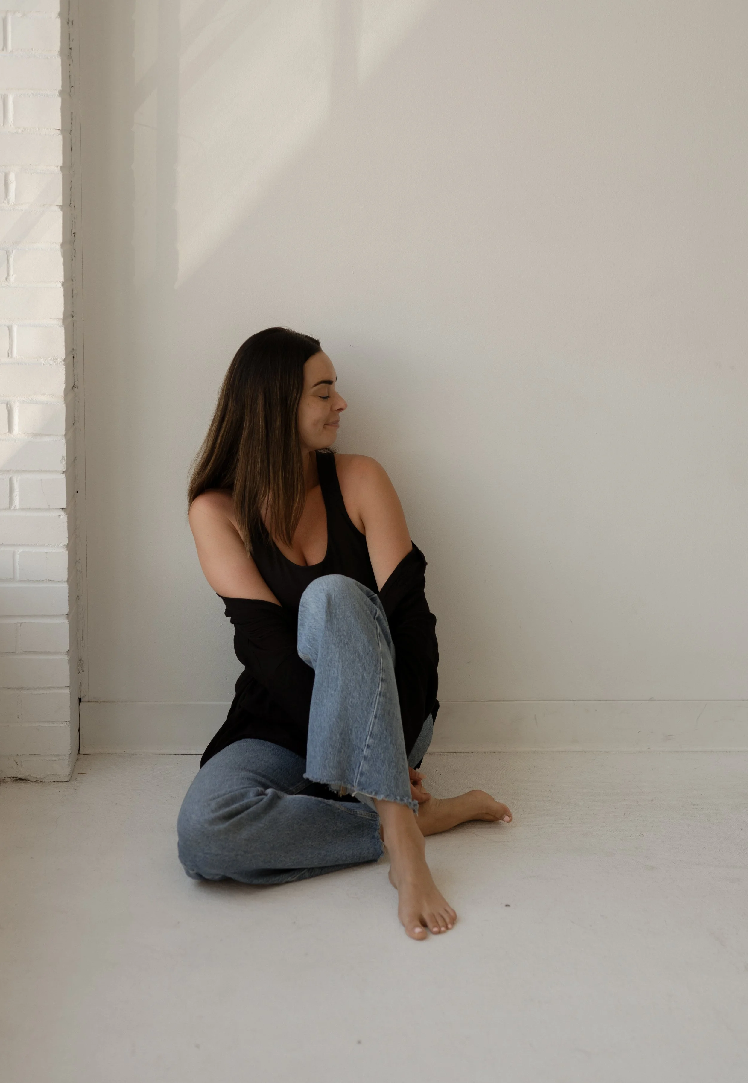 A woman with long brown hair, wearing a black tank top and gray sweatpants, sitting on the floor against a plain white wall with bare feet, smiling softly and looking to the side.