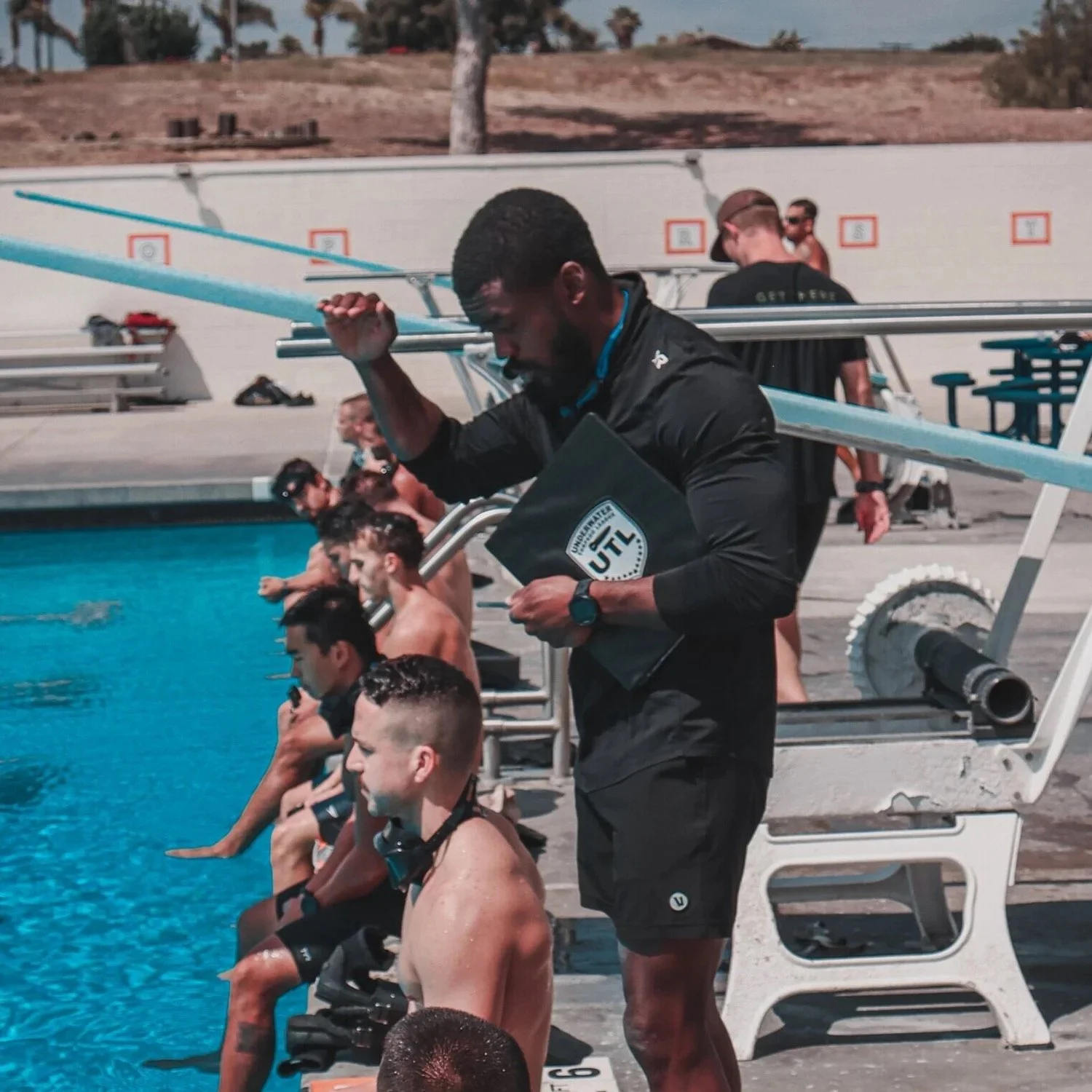 A swim coach with a clipboard stands at the edge of a swimming pool, instructing a line of young male swimmers seated on the poolside during a training session.