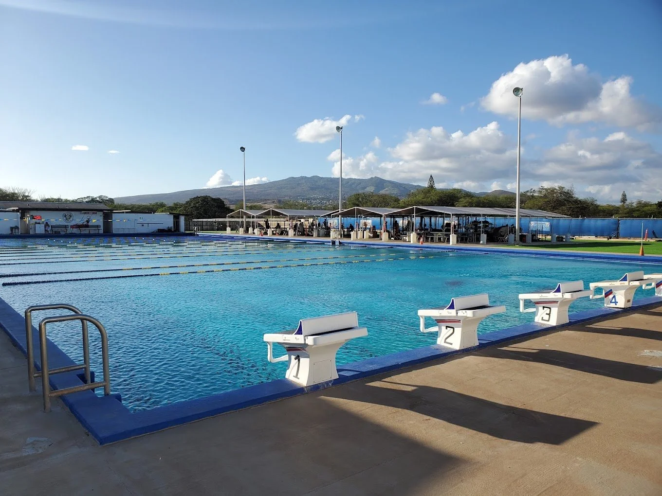 Outdoor swimming pool with clear blue water, surrounded by a concrete deck, with a green building, palm trees, and a bright blue sky in the background.