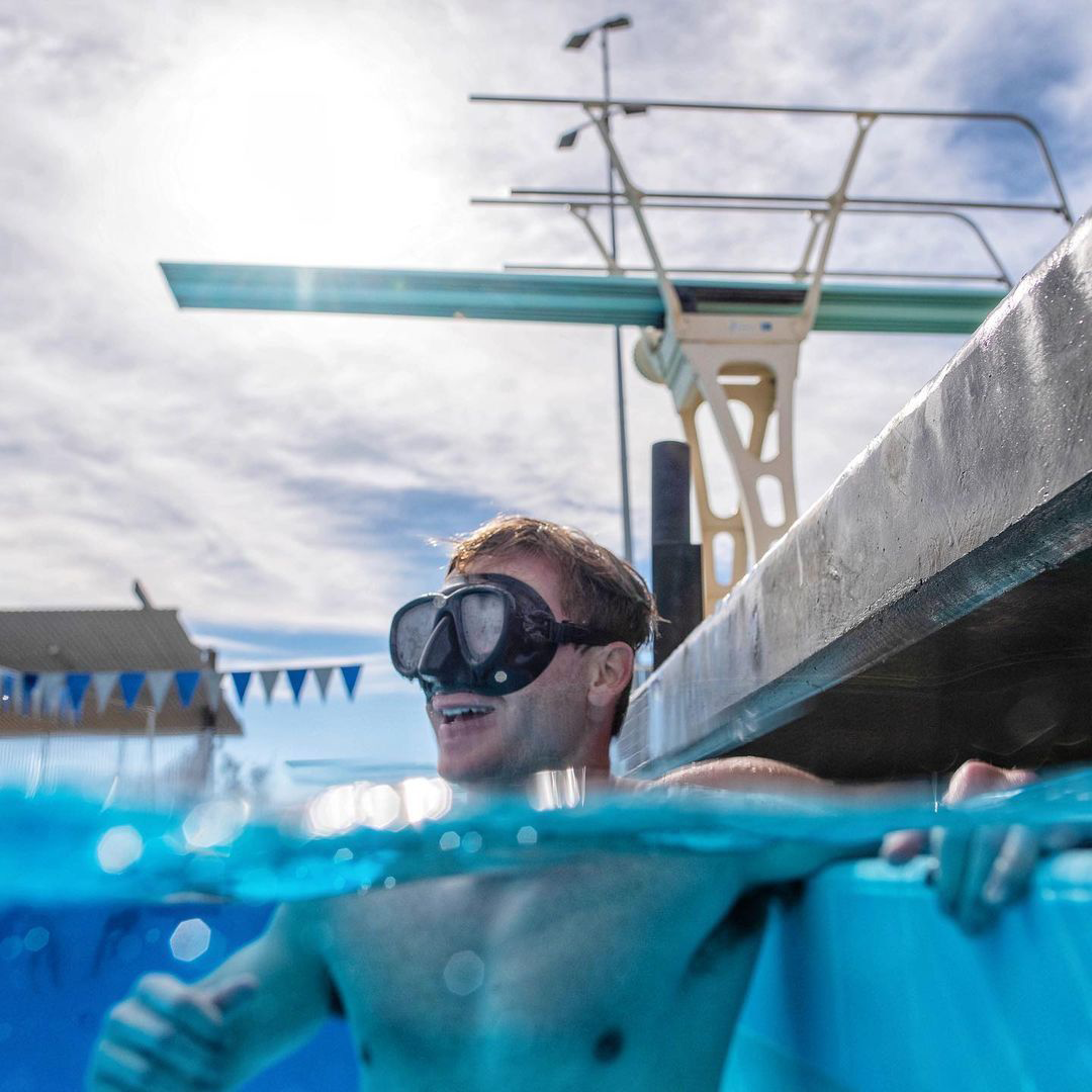 Man wearing swimming goggles smiling in a swimming pool, partly submerged with water level at his shoulders, with a diving board and sky in the background.