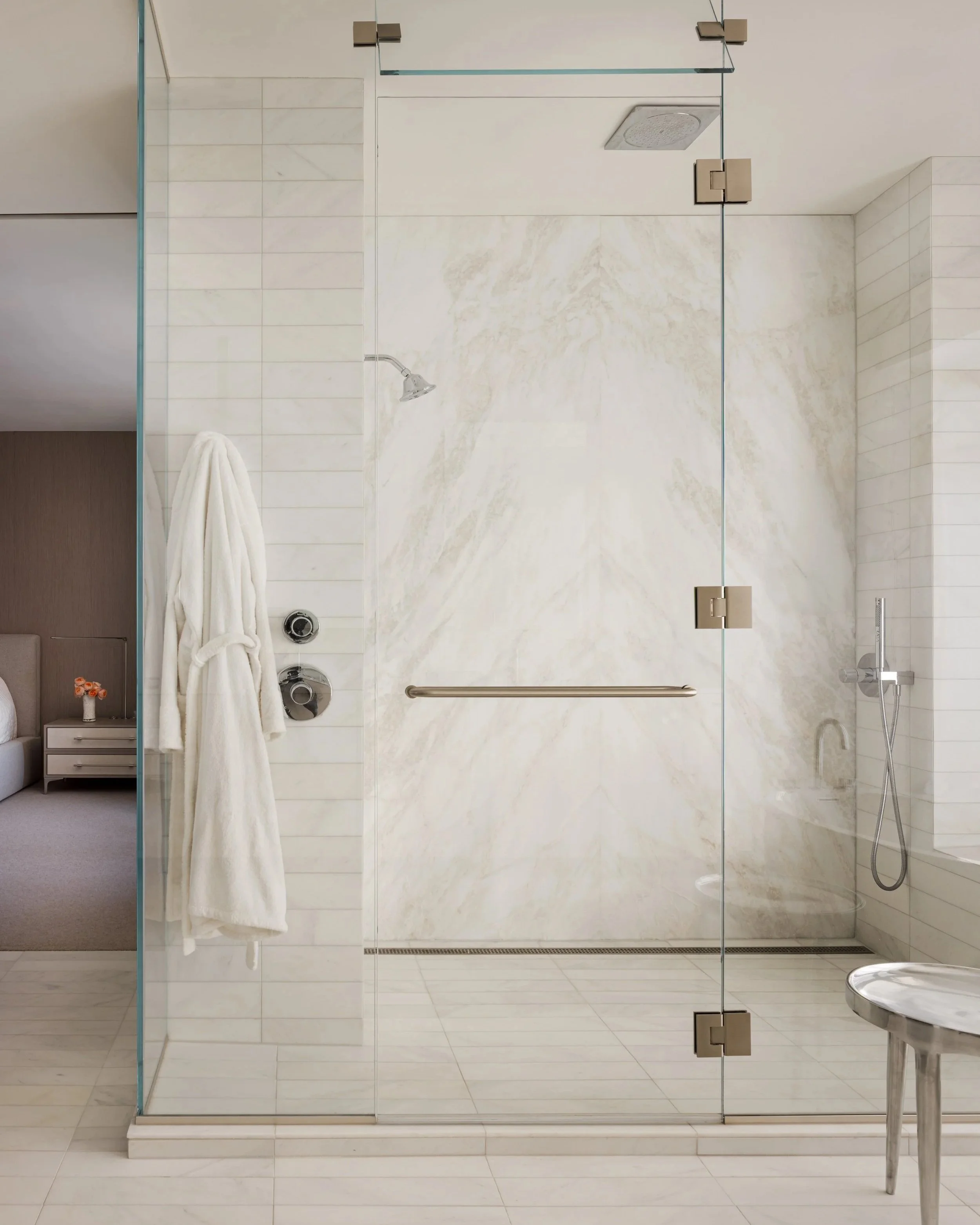 Modern glass-enclosed shower with marble walls, a handheld showerhead, a ceiling-mounted shower head, a towel, and a grab bar. Part of a bathroom with light-colored tiles and a seat outside the shower.