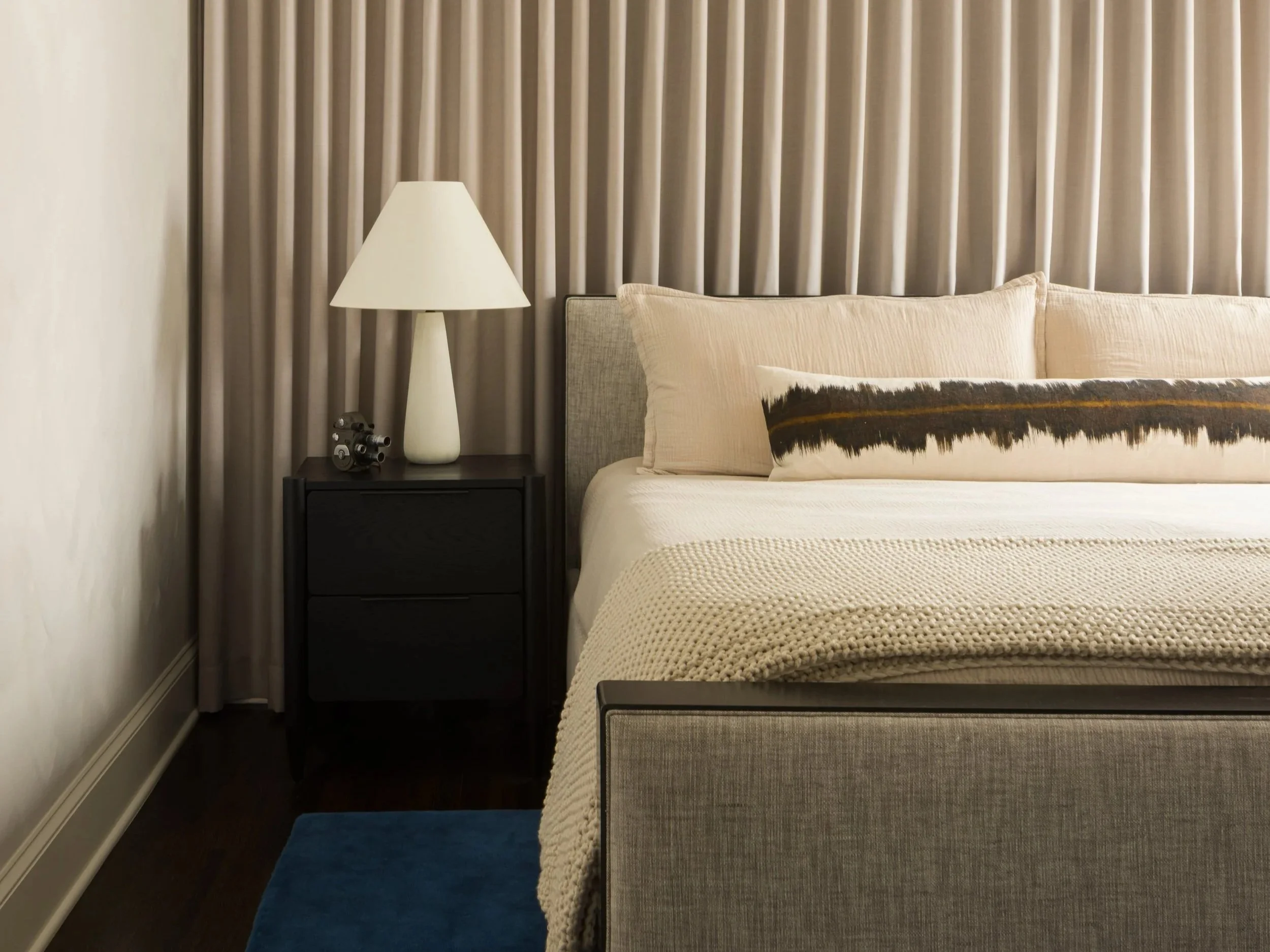 A bedroom with a bed, beige pillow, and horizontal decorative pillow, a bedside table with a white lamp, a small decorative clock, and beige curtains in the background.