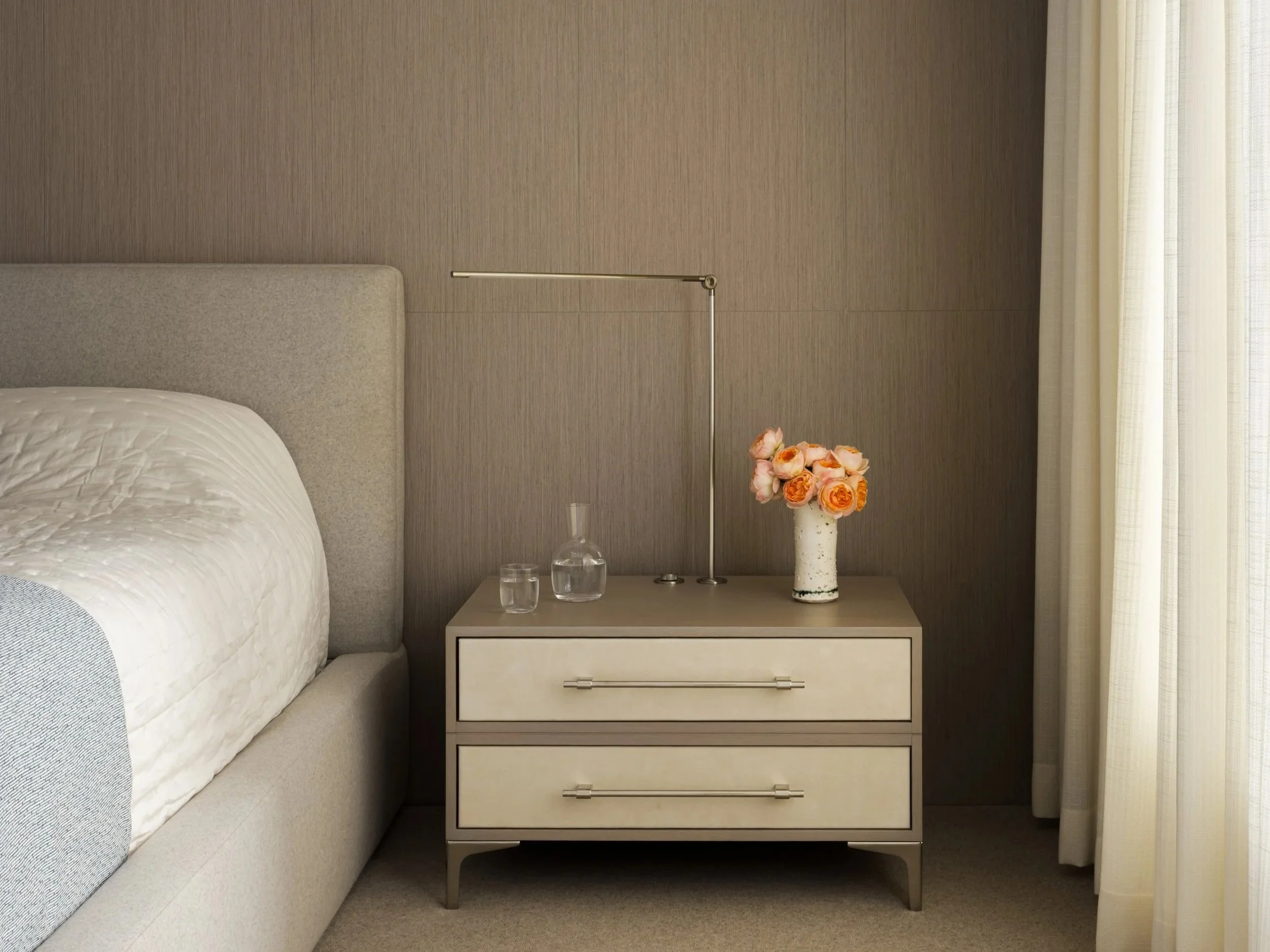 Bedroom nightstand with a vase of pink roses, a glass decanter, and a glass of water, next to a bed with a textured white comforter and a padded headboard, with beige curtains on the right.