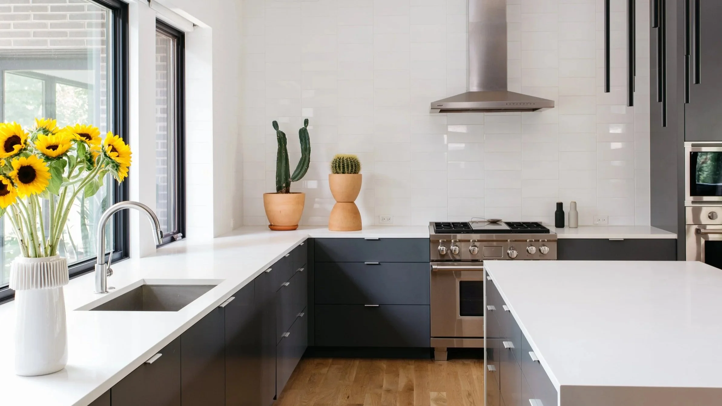 Modern kitchen with black cabinets, white countertops, and a large white tile backsplash. Sunflowers in a vase are on the left side near a window, along with potted cacti and a round cactus on the counter. Stainless steel appliances and black pendant
