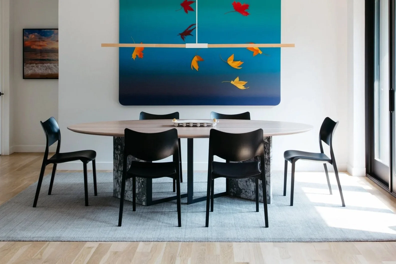 Modern dining room with round wooden table, six black chairs, large artwork with falling leaves on the wall, and sliding glass doors on the right.