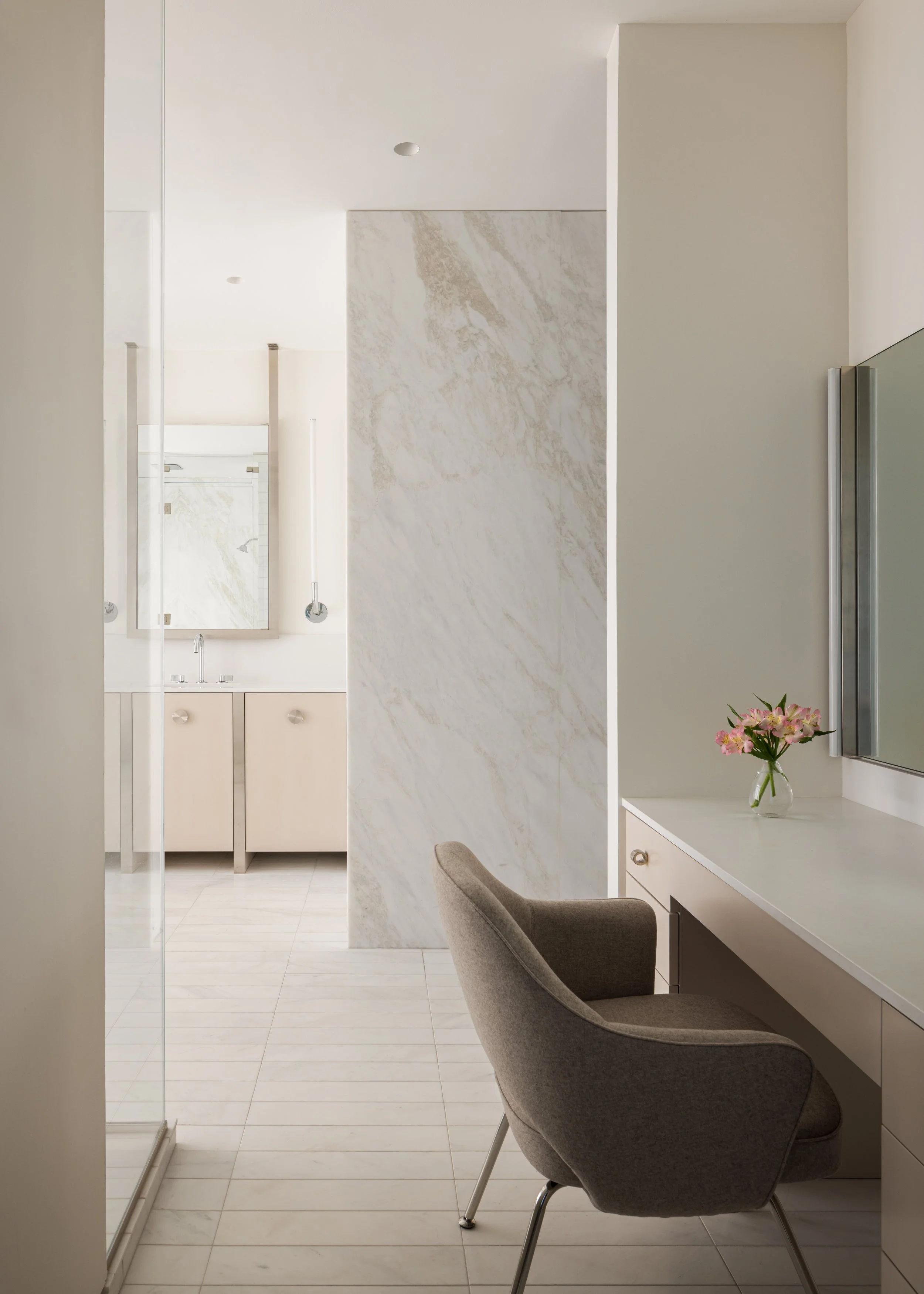 A modern, minimalistic bathroom with a neutral color palette, featuring a vanity with a mirror, a vase with pink flowers on the desk, and a cozy chair nearby.