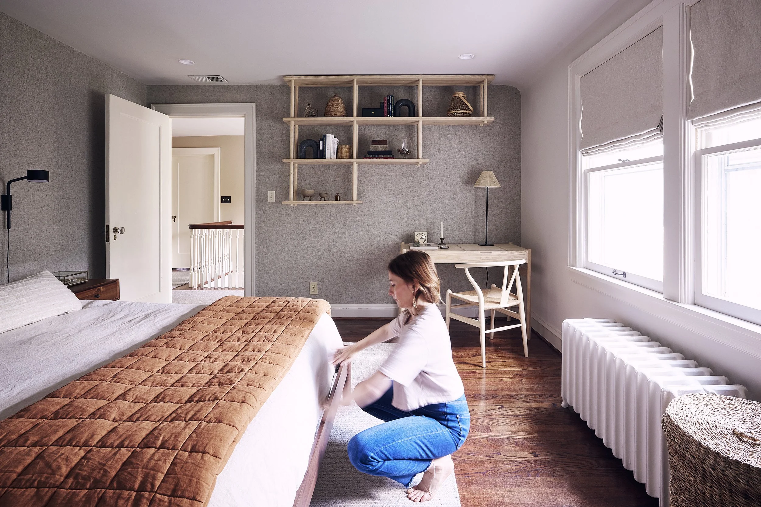 A woman kneeling on the floor, adjusting a brown quilt on a bed in a bright, modern bedroom with large windows and a wooden floor.