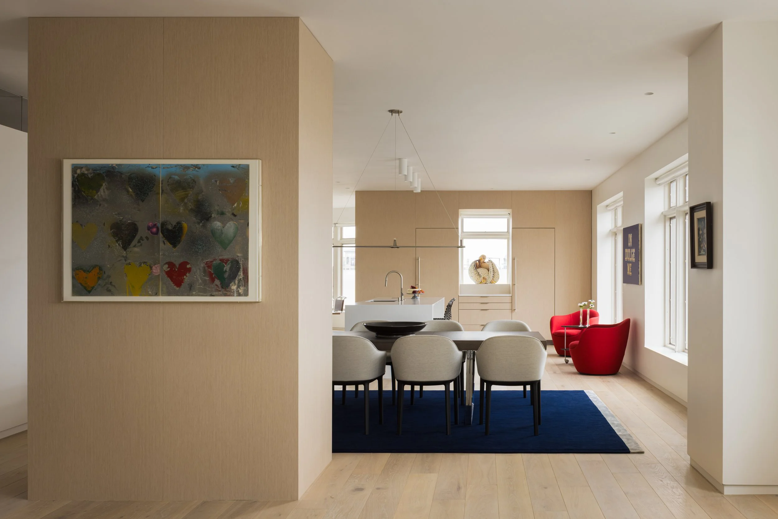 Open-plan dining and living area with beige walls, a dining table with chairs, and a sitting area with red chairs near windows.