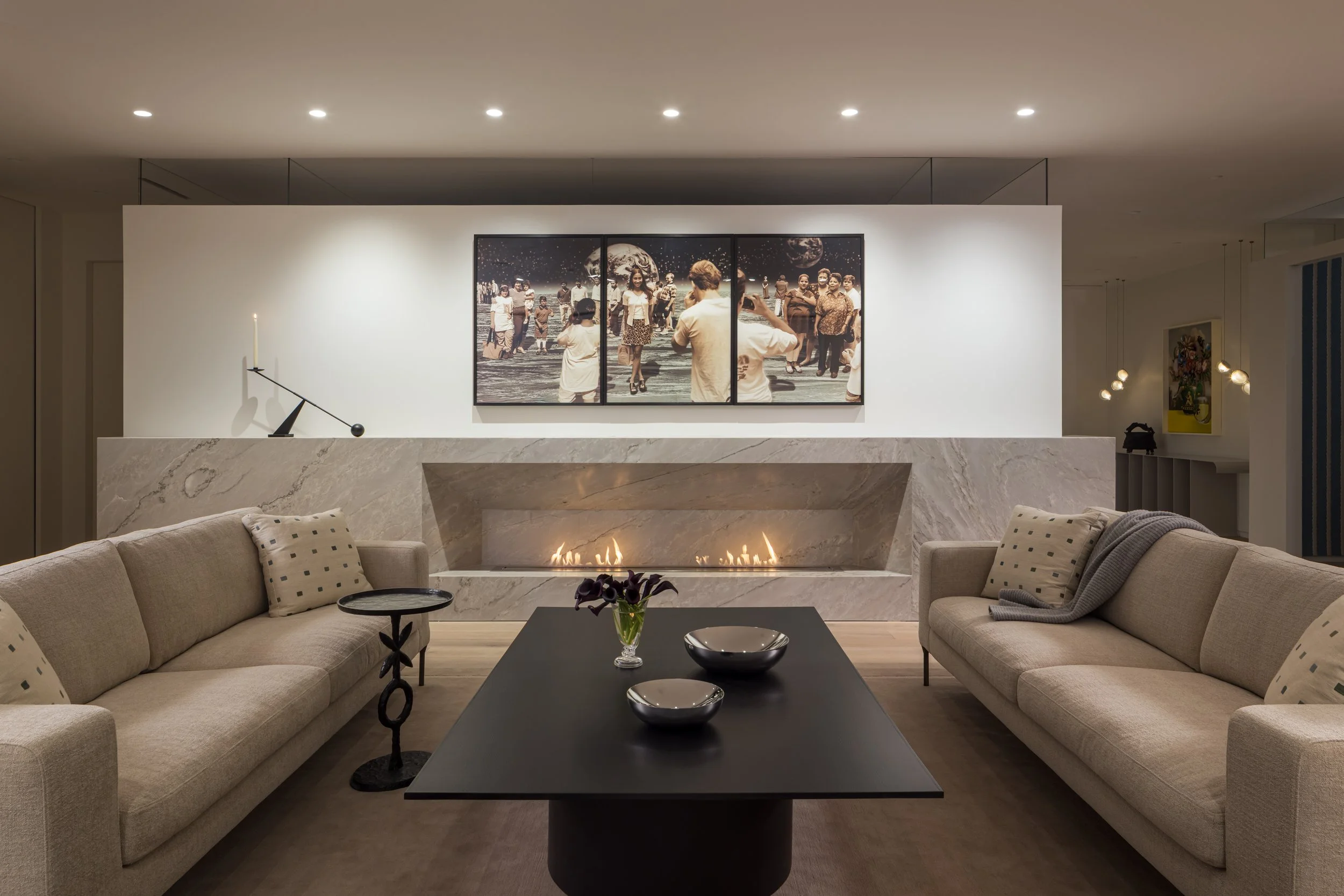 Living room with a modern fireplace, beige sofas, a black coffee table with bowls and flowers, and a large multi-panel wall art depicting people on the moon.