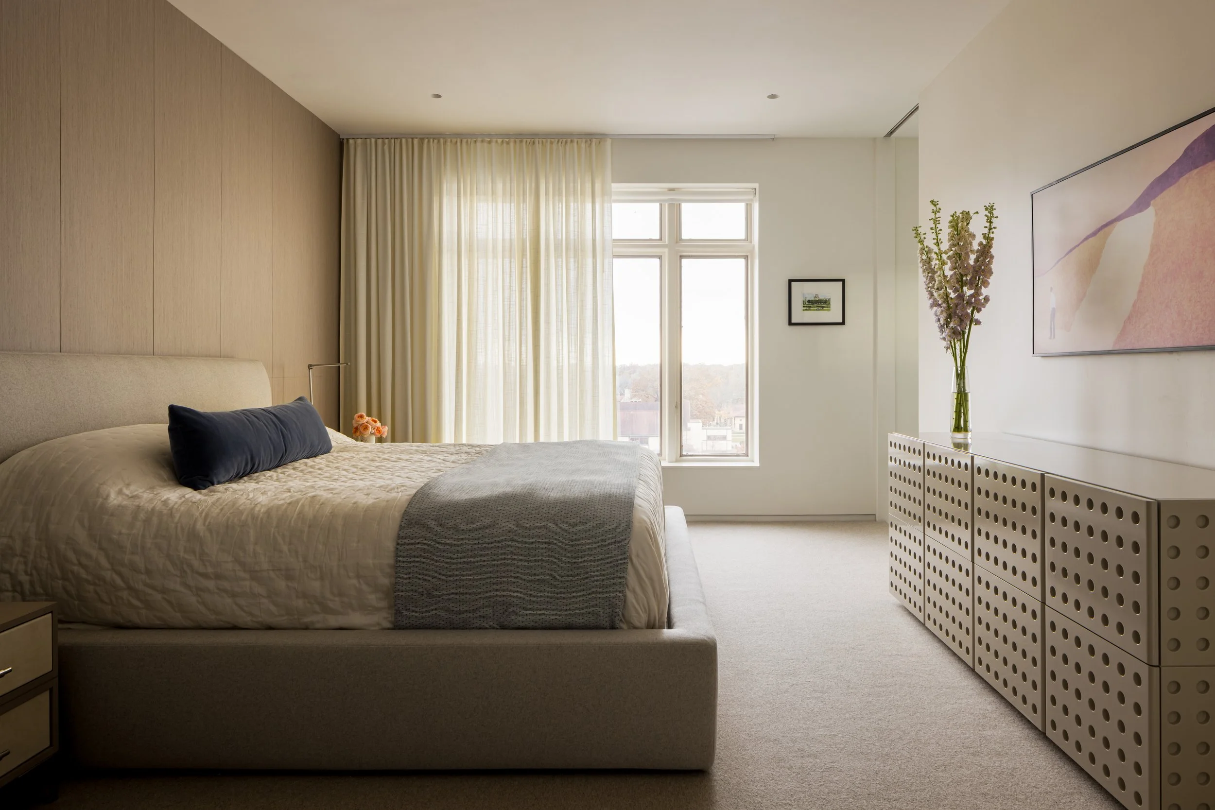 A bedroom with a large bed, beige headboard, navy blue pillow, beige quilt, and gray blanket. Light from a window with cream curtains illuminates the room. There is a small picture on the wall, a tall vase with flowers on a white cabinet, and Abstrac