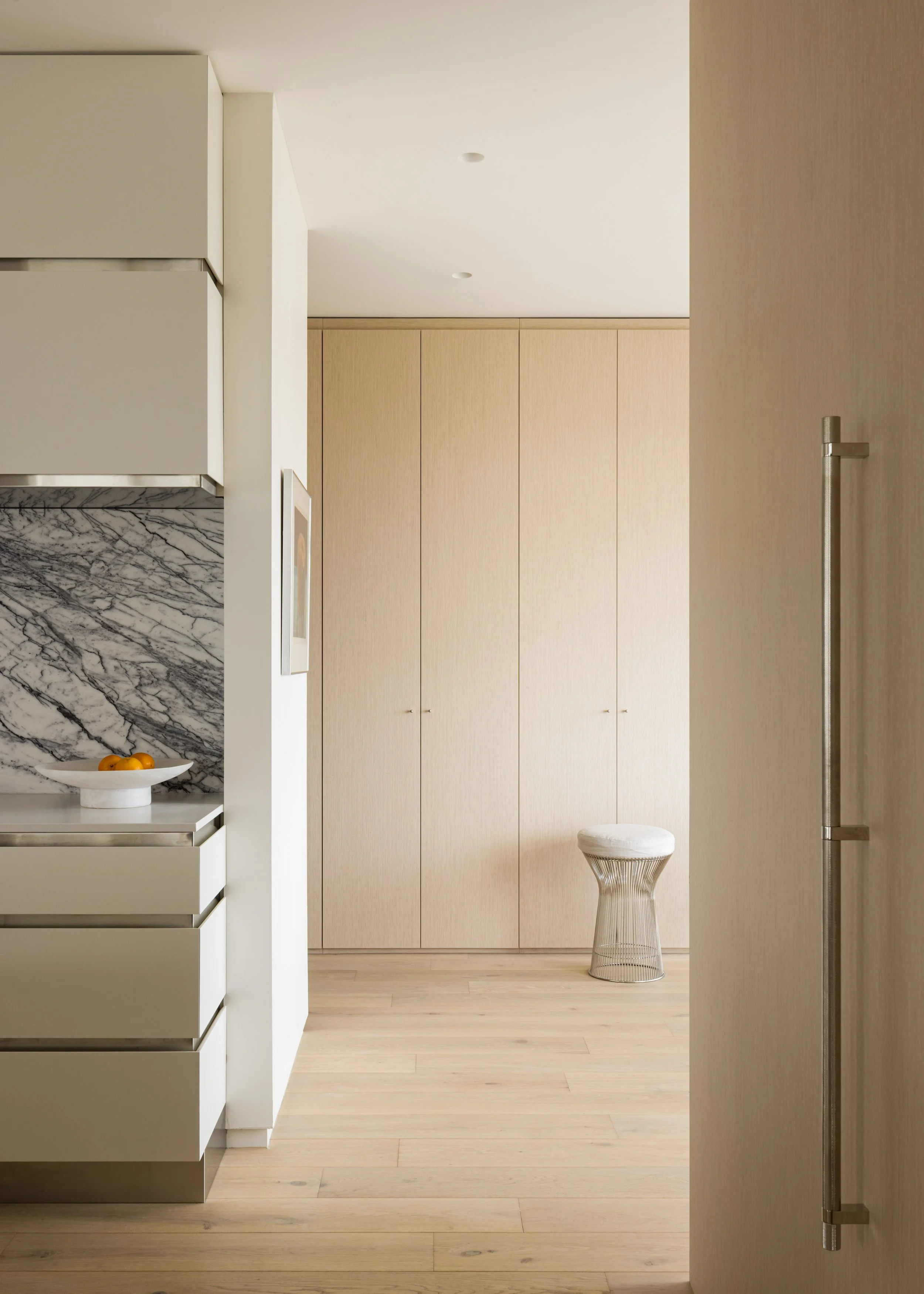 Minimalist kitchen with bleached white oak wooden cabinets and a Vernon Platner Collection Wire Frame upholstered stool with white cushion. Stainless steel band details between cabinet drawers add sophistication. 