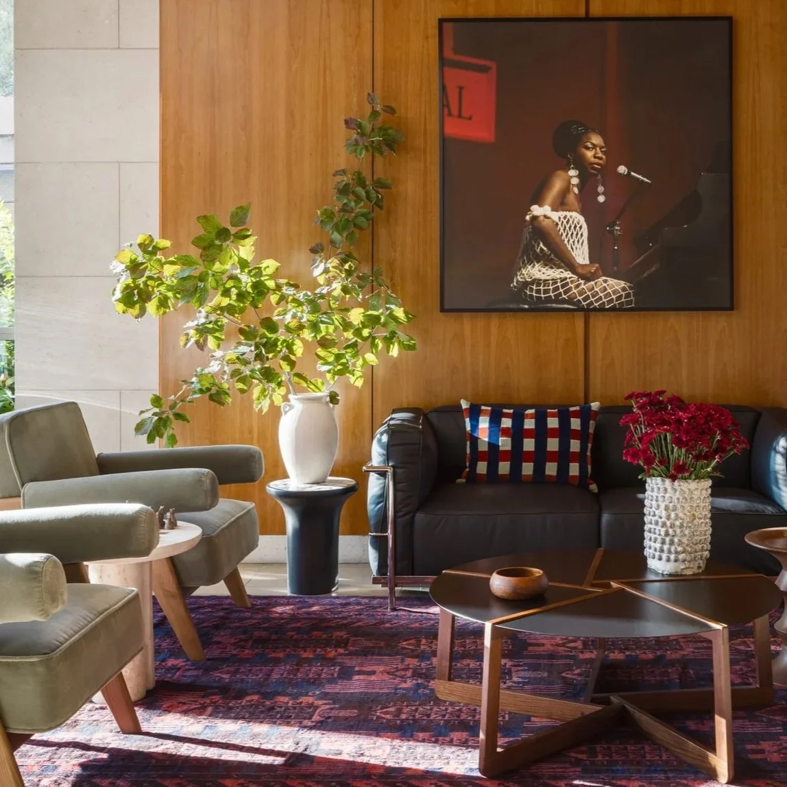 A cozy living room with green armchairs, a black sofa, a side table, and a wooden coffee table with a vase of red flowers, a large plant in a white vase, framed photograph of a woman at a piano on the wooden wall, and a colorful patterned rug.