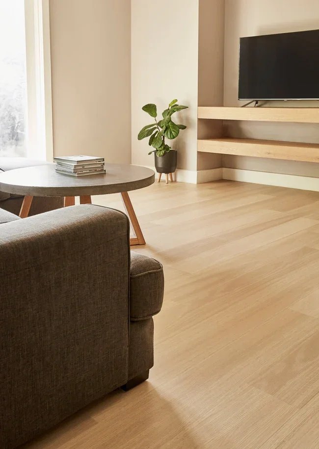 A spacious, modern lounge area with large floor-to-ceiling windows showing a city skyline. The room features Pink, white, and blue cushioned ottomans scattered around a gray patterned carpet. There's a white shelf with books and decorative items on the wall, and a small green plant in the corner.
