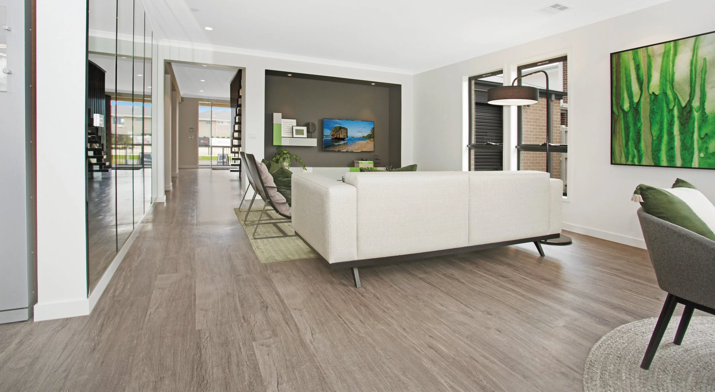 Modern living room with white sofa, black chairs, wall-mounted TV, green accents, large windows, wooden flooring, and artwork on the wall.