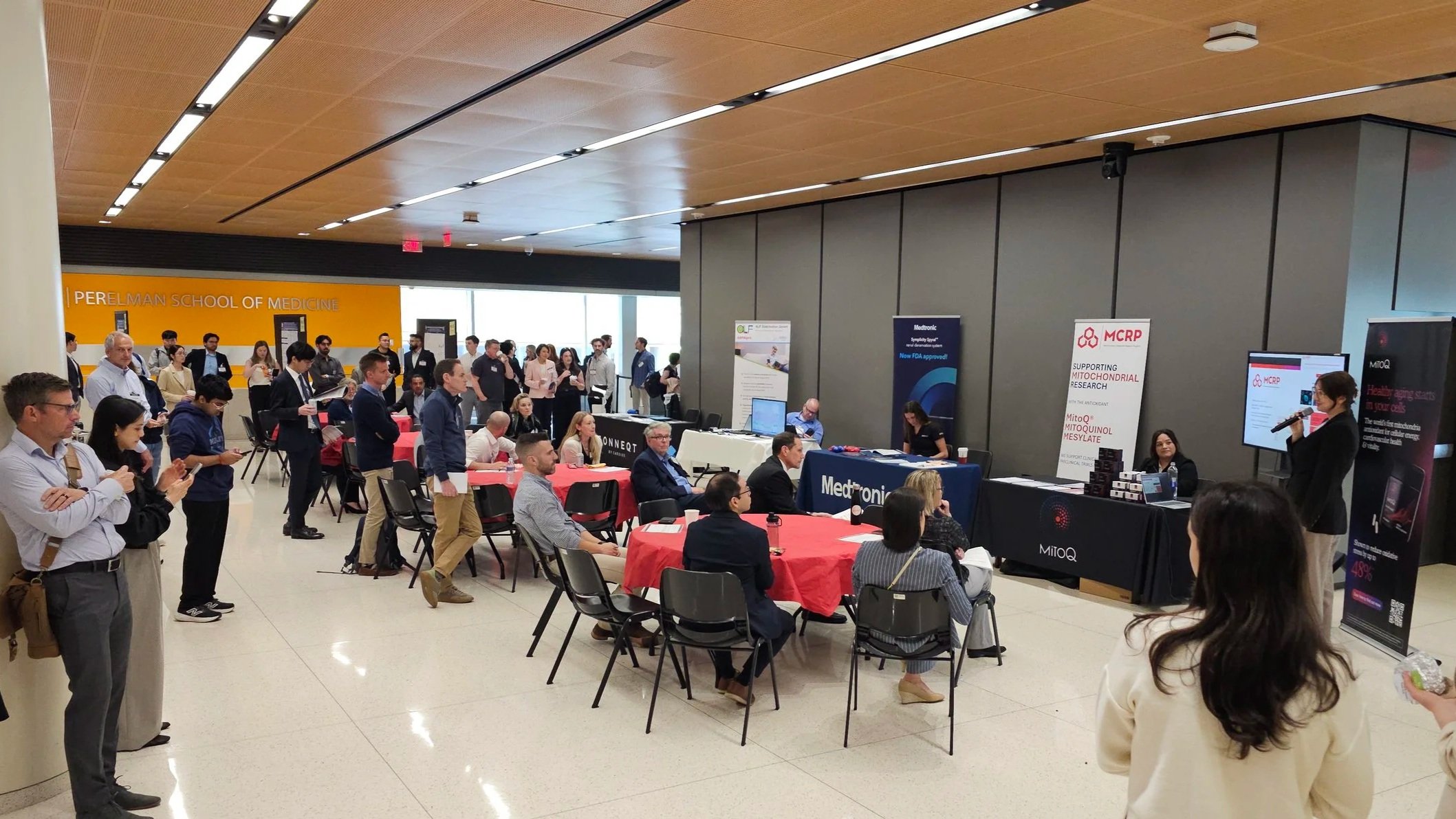 A seminar on Mitoquinol taking place at the Perelman School of Medicine with attendees and exhibitors, some engaging with displays and presentations.