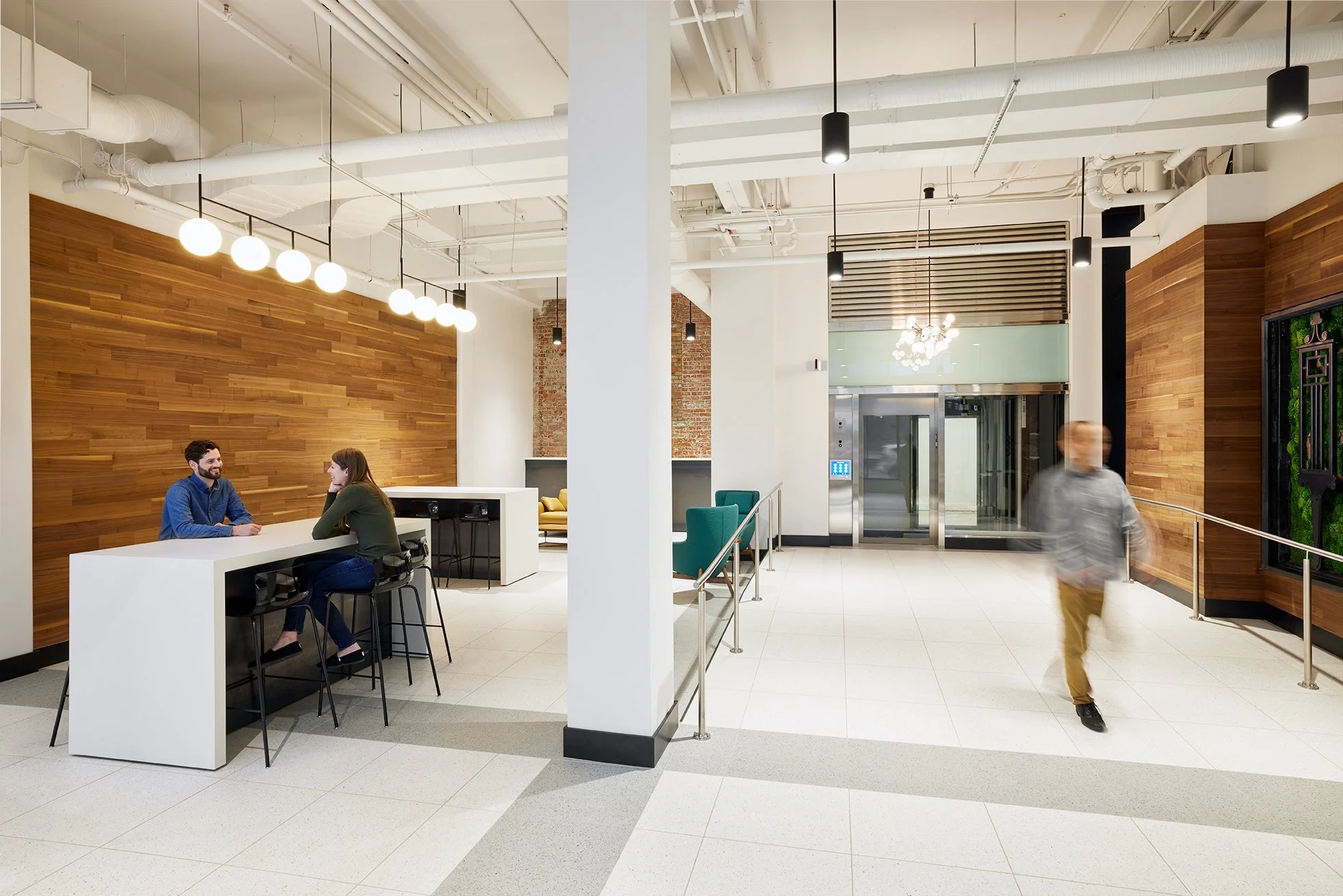 Architectural Modern design of an office lobby with wooden wall panels, white walls, and tile floors. Two people seated at a tall white tables engaged in conversation. A blurred person walking across the lobby near elevators.