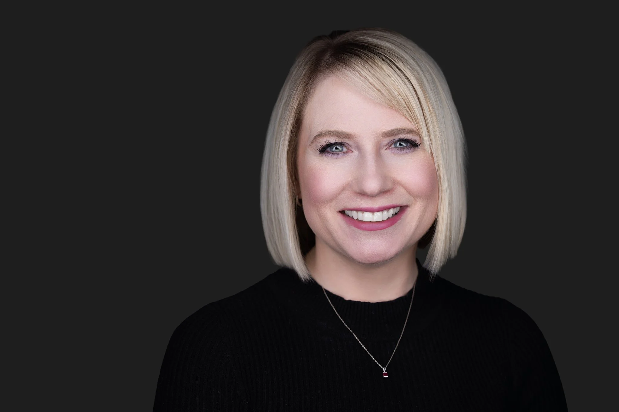 Headshot of a woman with shoulder-length blonde hair, smiling and wearing a black top against a dark background showing depth 