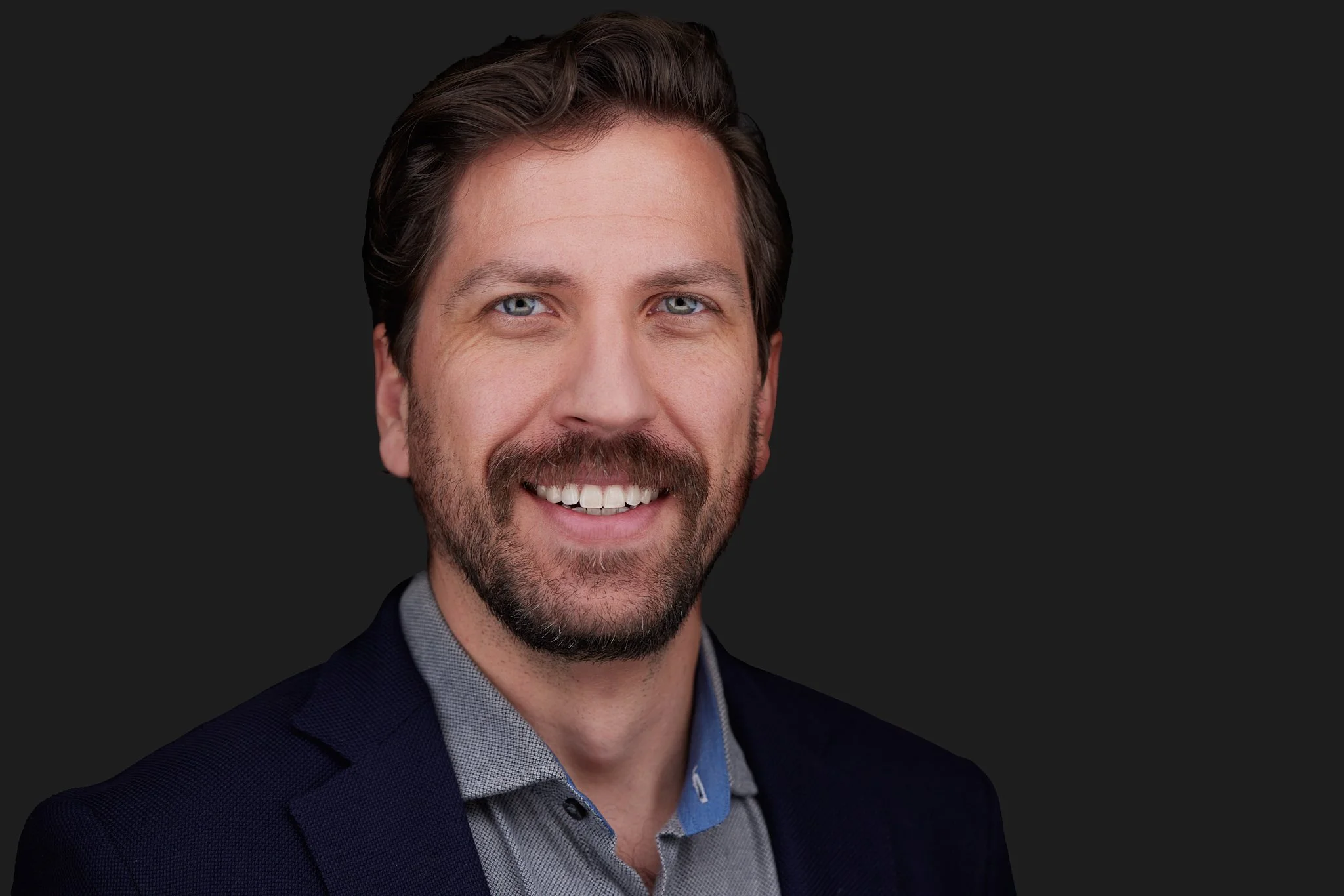 Team Headshot of a smiling man wearing a dark blazer and patterned shirt, against a black background.