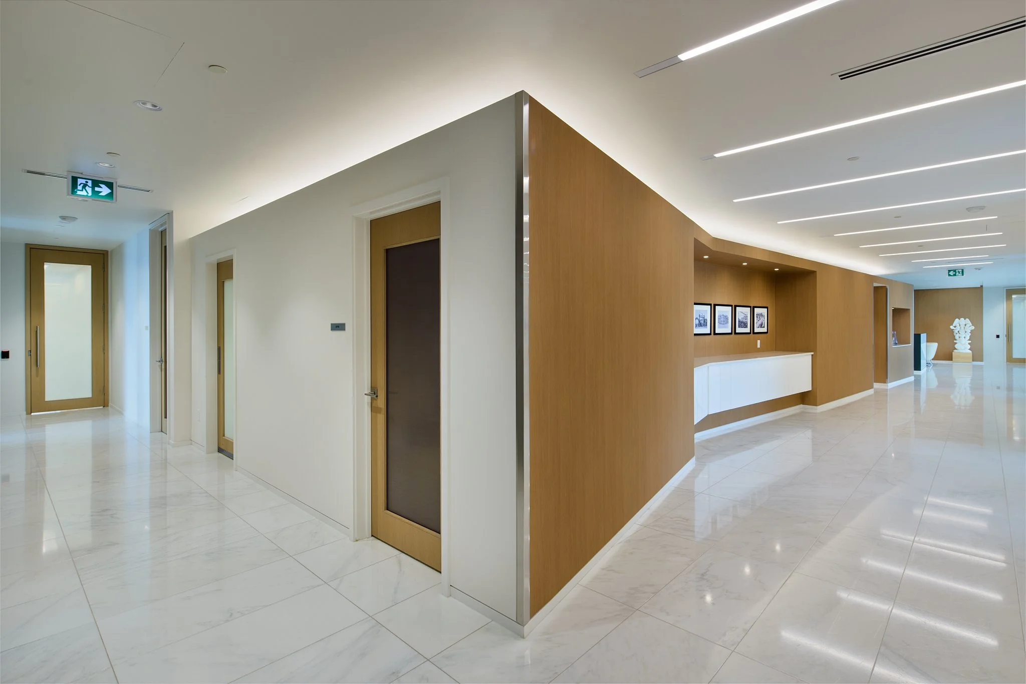 Modern interior hallway with marble flooring, white and wood-paneled walls, framed black-and-white pictures, and ceiling with linear lighting.
