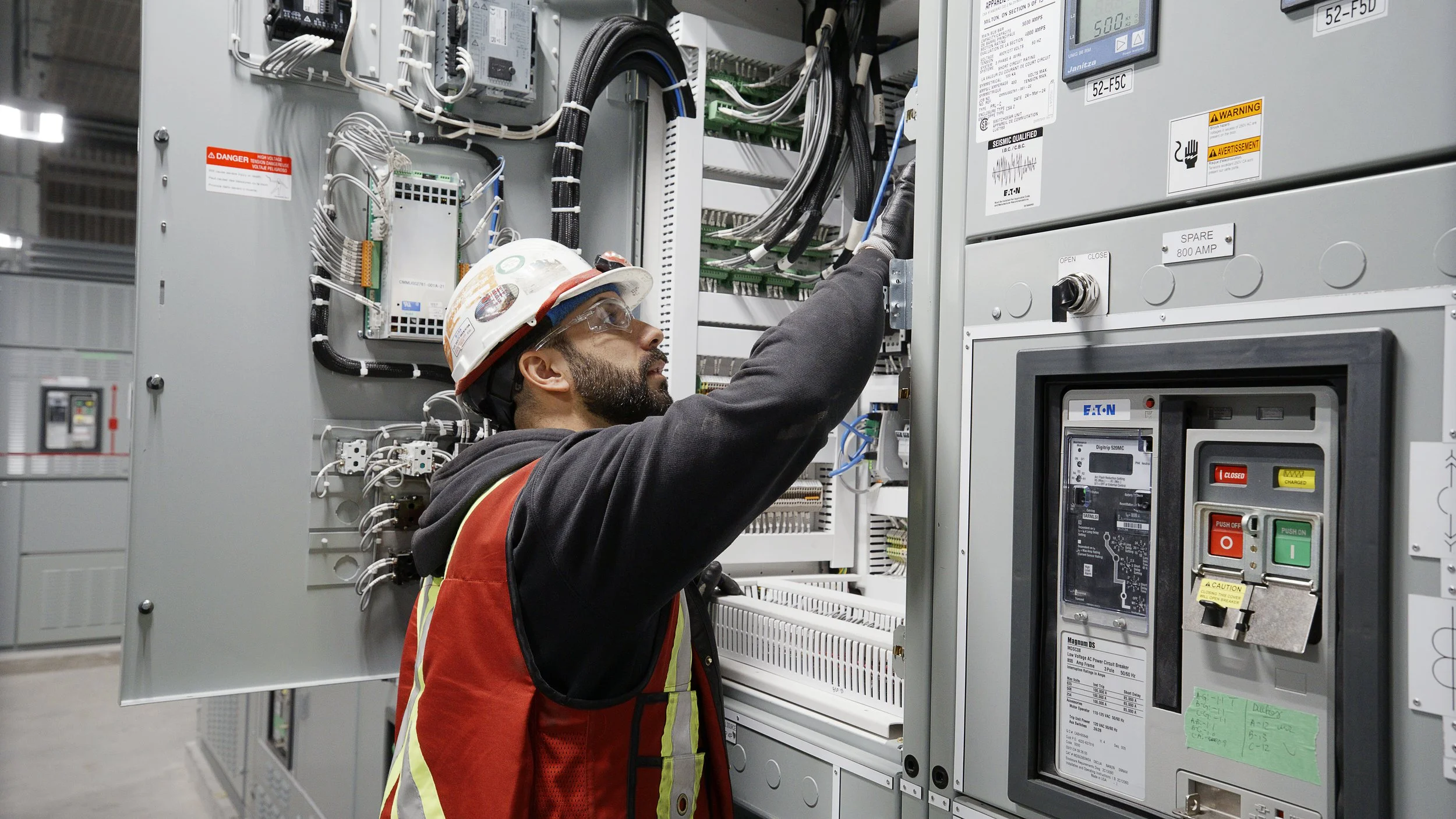 A worker wearing safety glasses and a helmet inspects electrical controls inside a large industrial electrical panel.