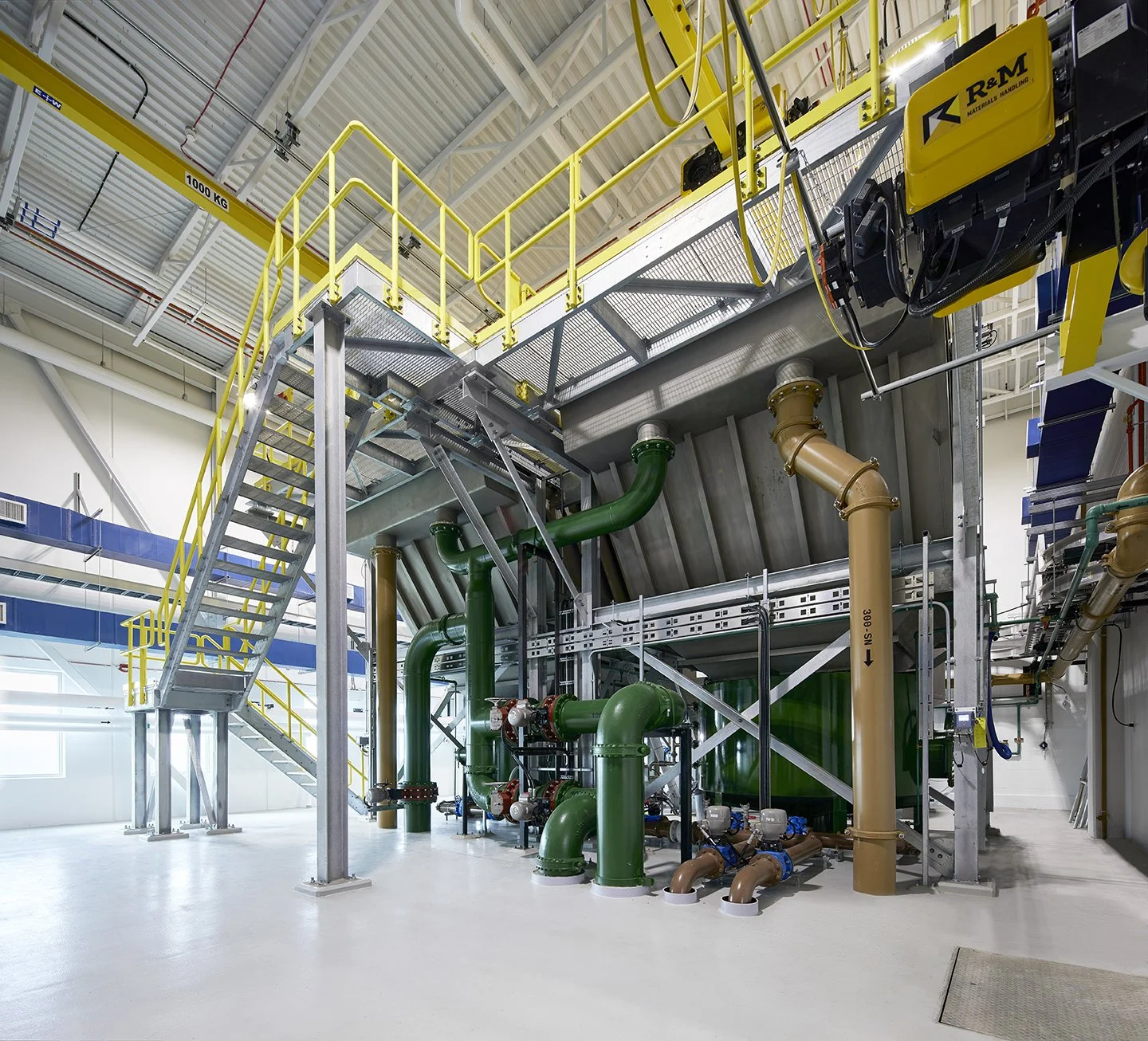 Industrial mechanical room with green and brown pipes, a metal staircase with yellow safety railings, and various machinery at an industrial facility.