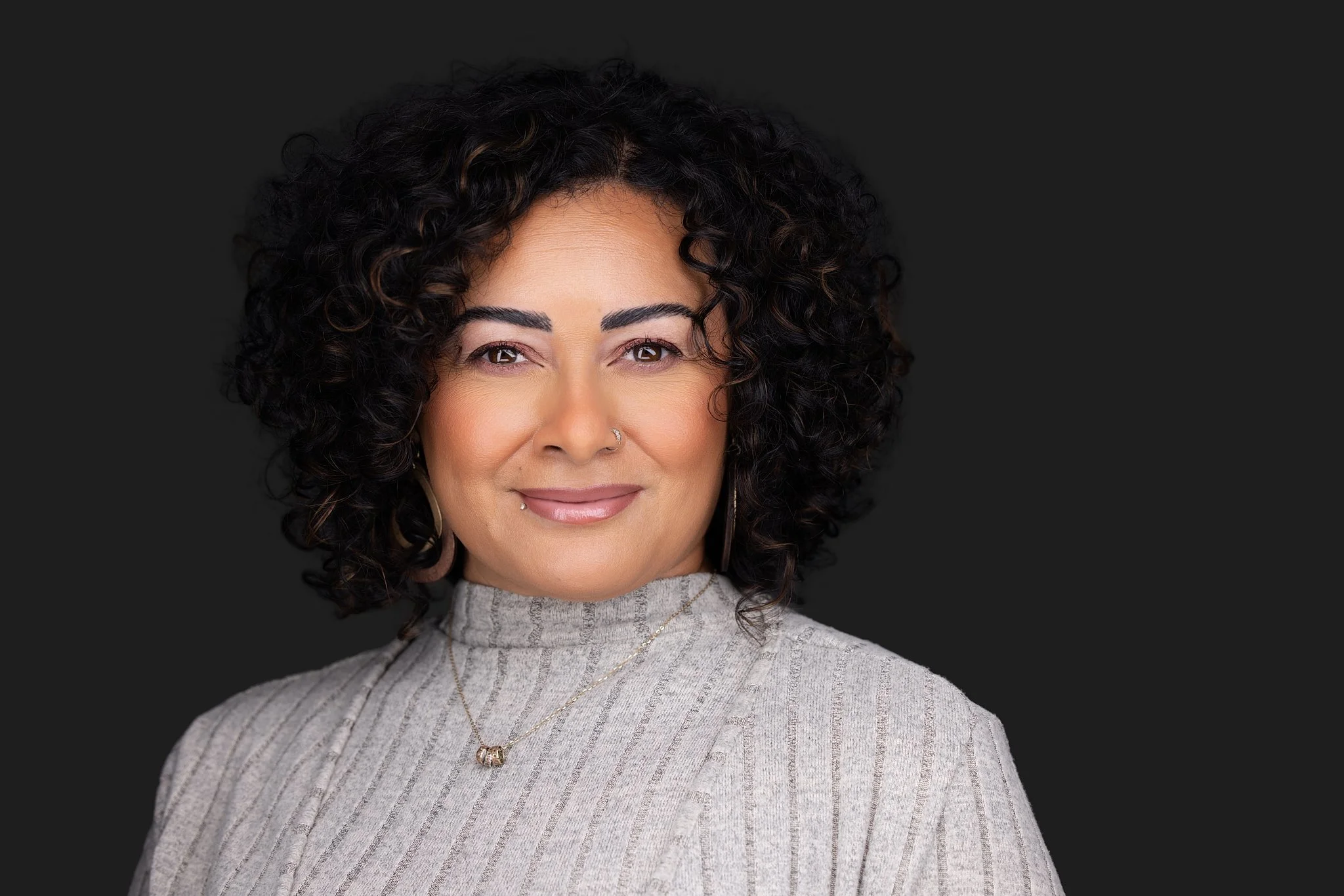 Corproate headshot of a woman with dark, curly hair smiling against a dark background.