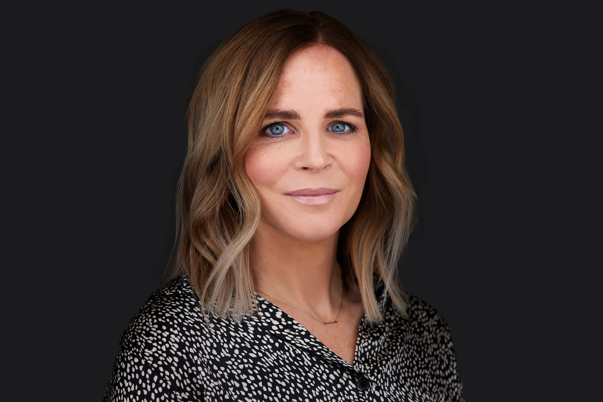 Corporate Headshot of a woman with shoulder-length wavy blonde hair, blue eyes against a dark background.