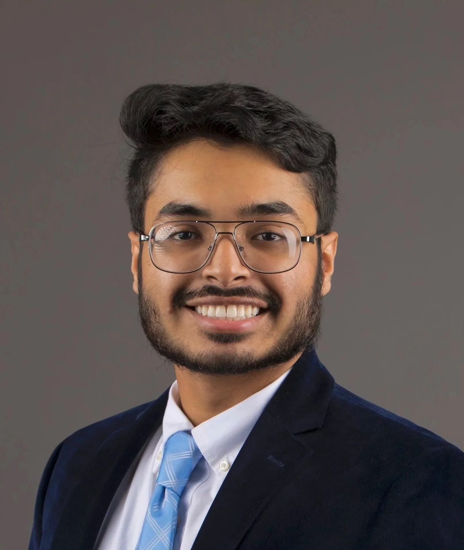 Professional headshot of a young man with glasses, dark hair, a beard, wearing a navy suit, white shirt, and light blue tie, smiling against a gray background.