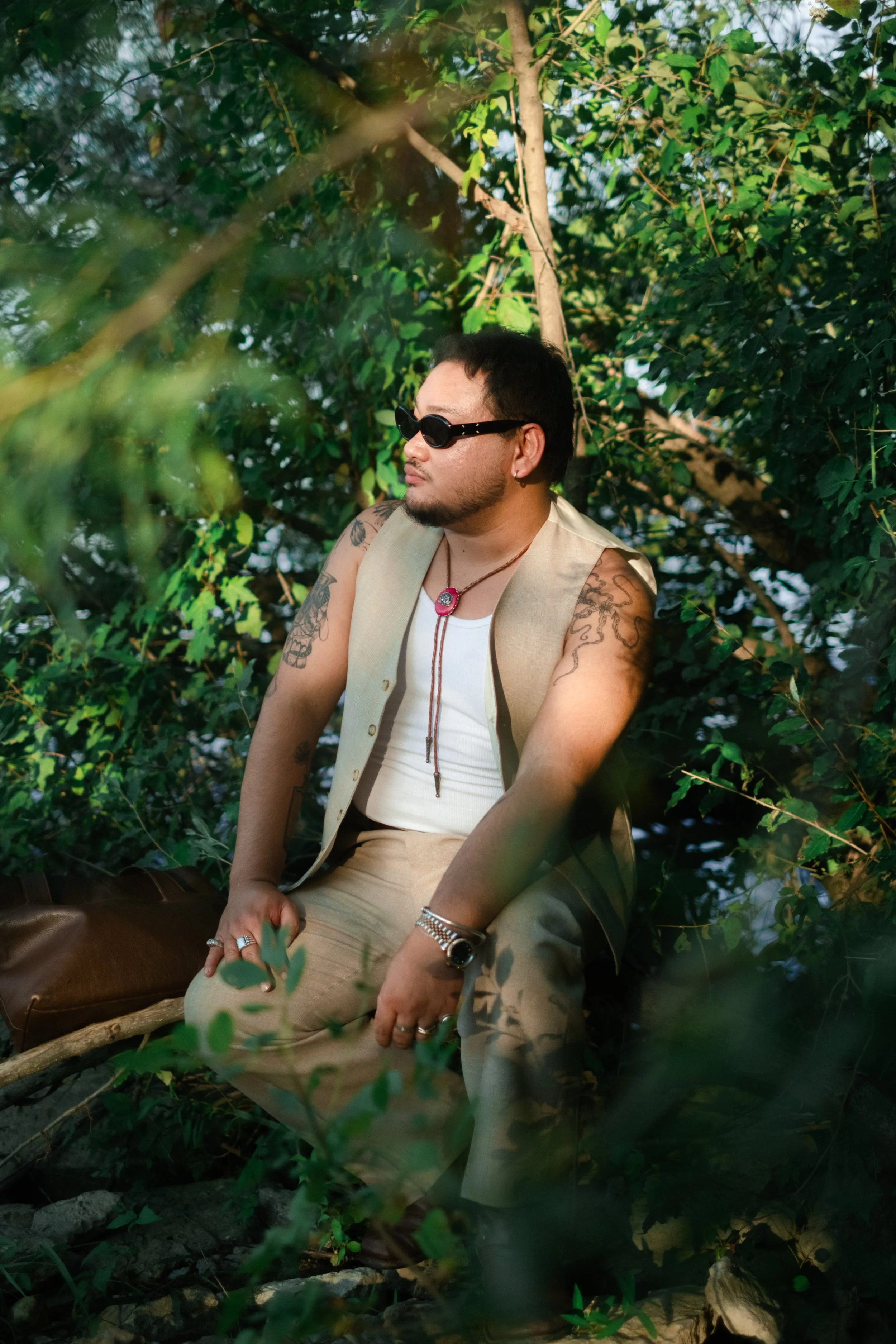 A man sitting outdoors in a lush, green setting, wearing sunglasses, a white sleeveless shirt, a beige vest, and camouflage pants, with tattoos on his arms, surrounded by leafy trees.