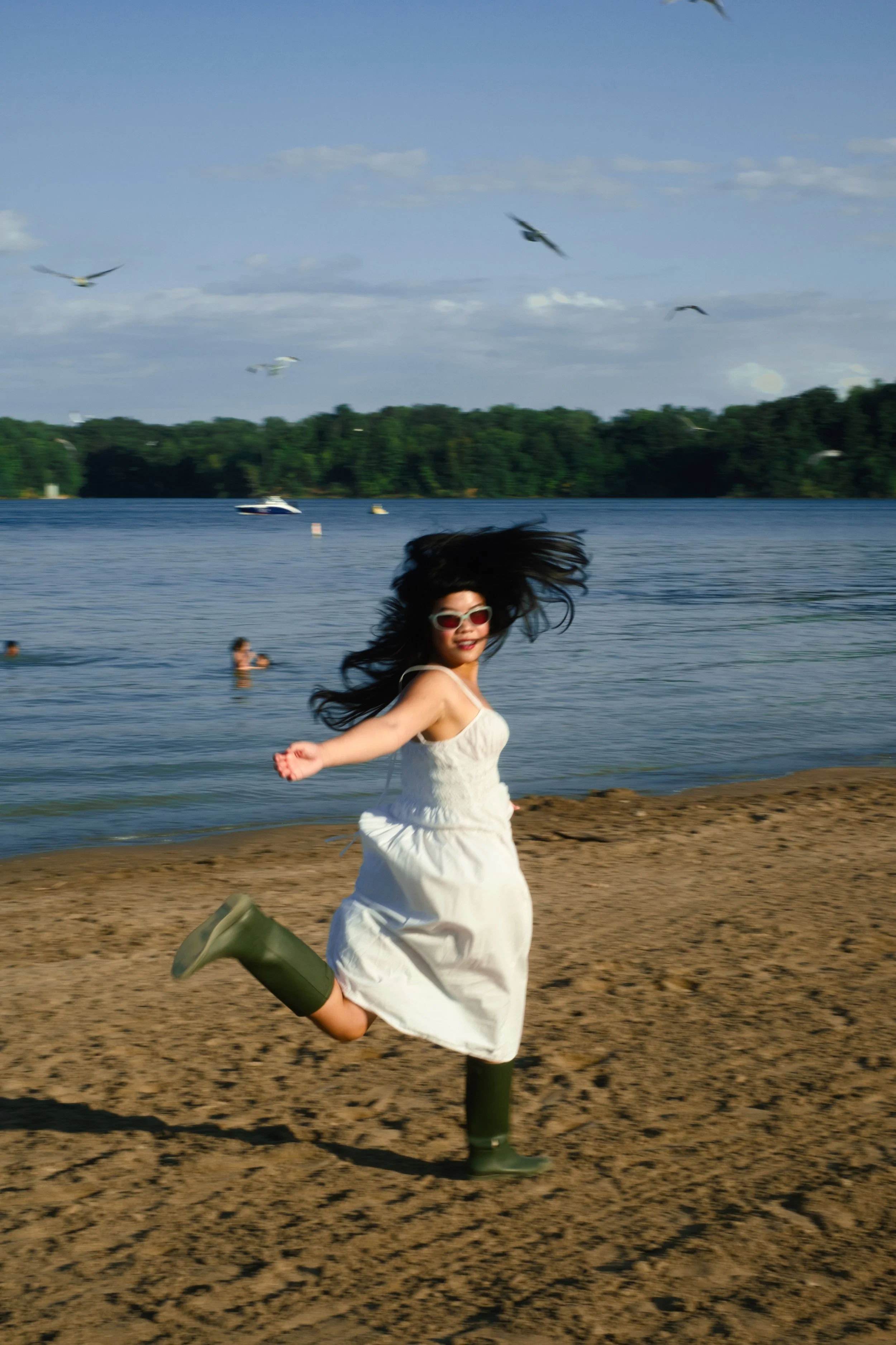 A woman running and smiling on a beach with water, boats, trees, and birds in the background.