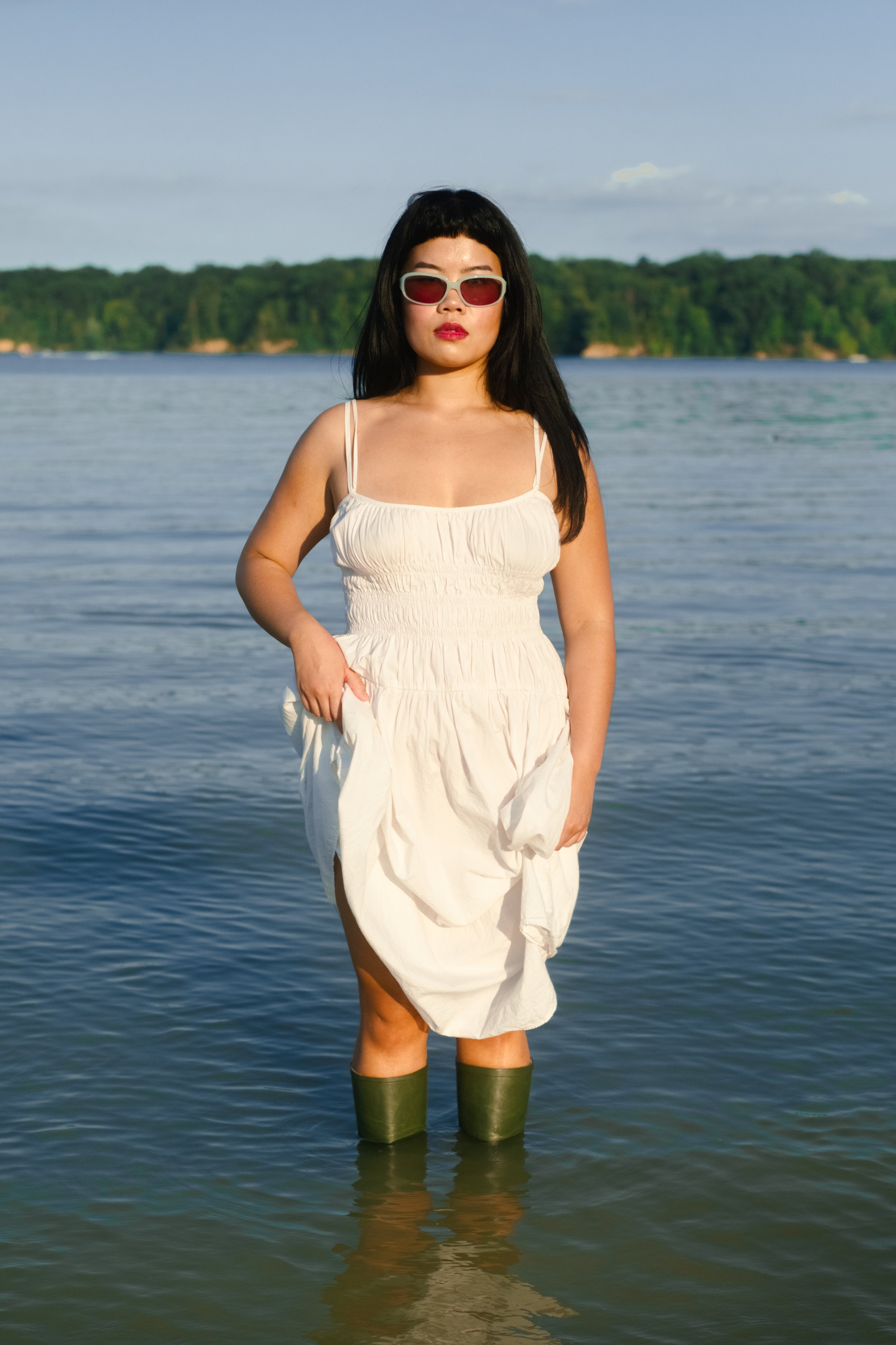 A woman wearing sunglasses and a white dress standing in water with a forested shoreline in the background.