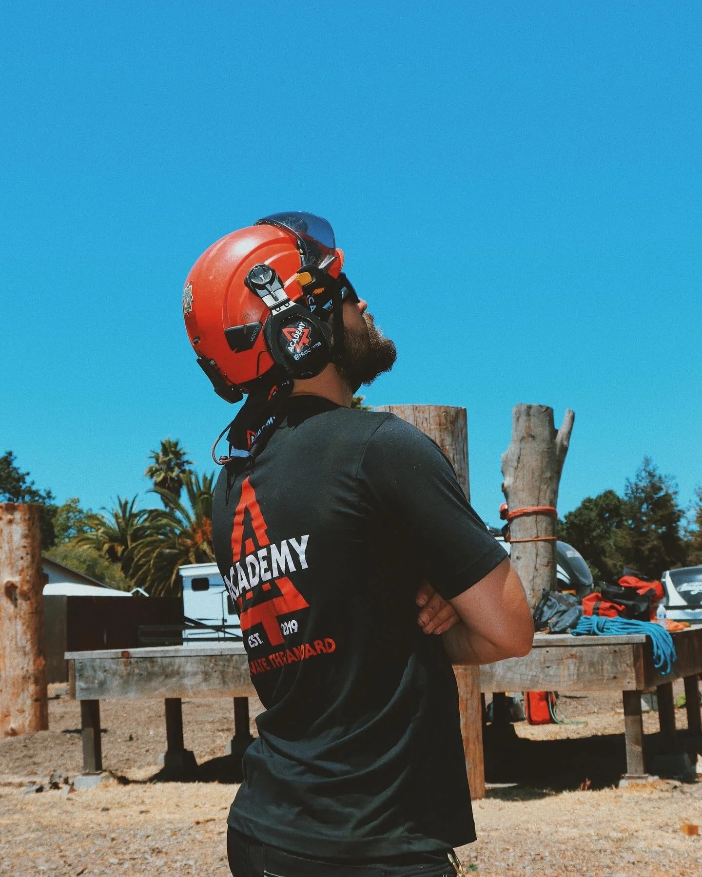 A man wearing a red safety helmet with a face shield and headphones, standing outdoors with arms crossed. In the background, there are trees, a wooden platform with tools and equipment, blue sky, and a few parked cars.