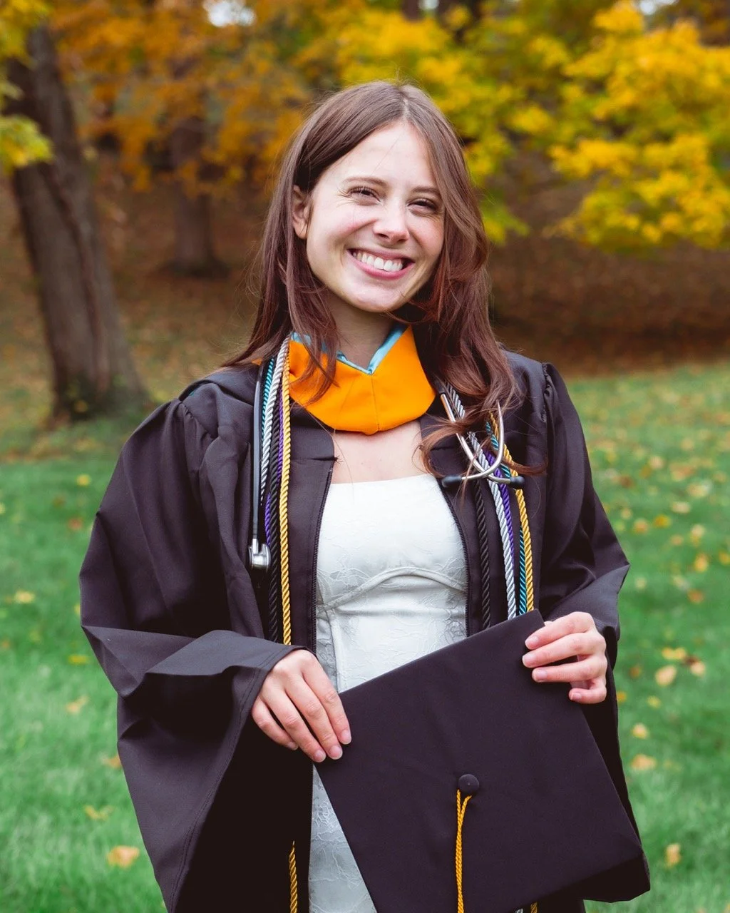A young woman in graduation regalia holding a diploma outdoors with autumn trees in the background.