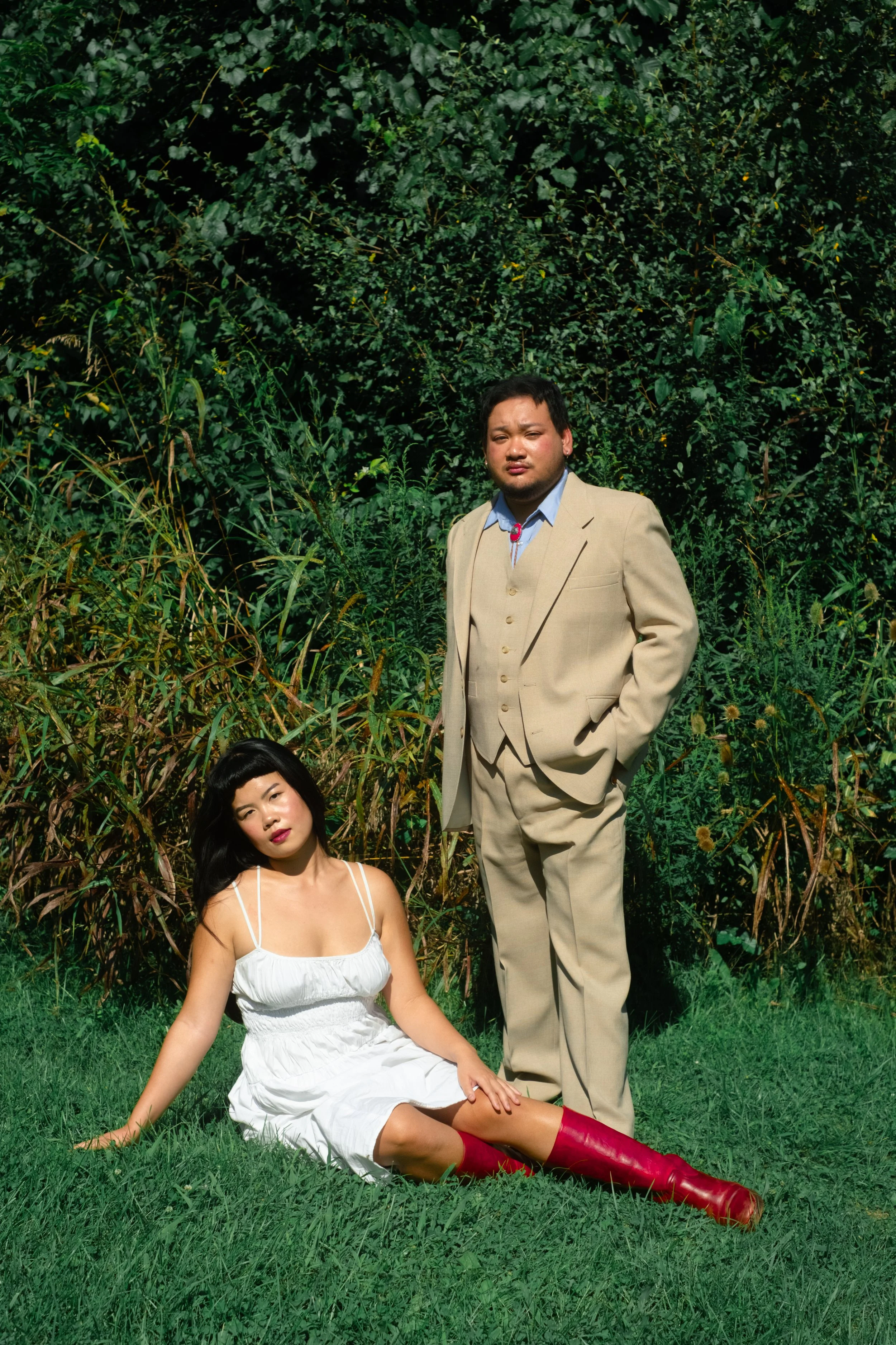 A woman in a white dress with red boots sitting on grass, a man in a beige suit standing behind her, both outdoors with green foliage in the background.