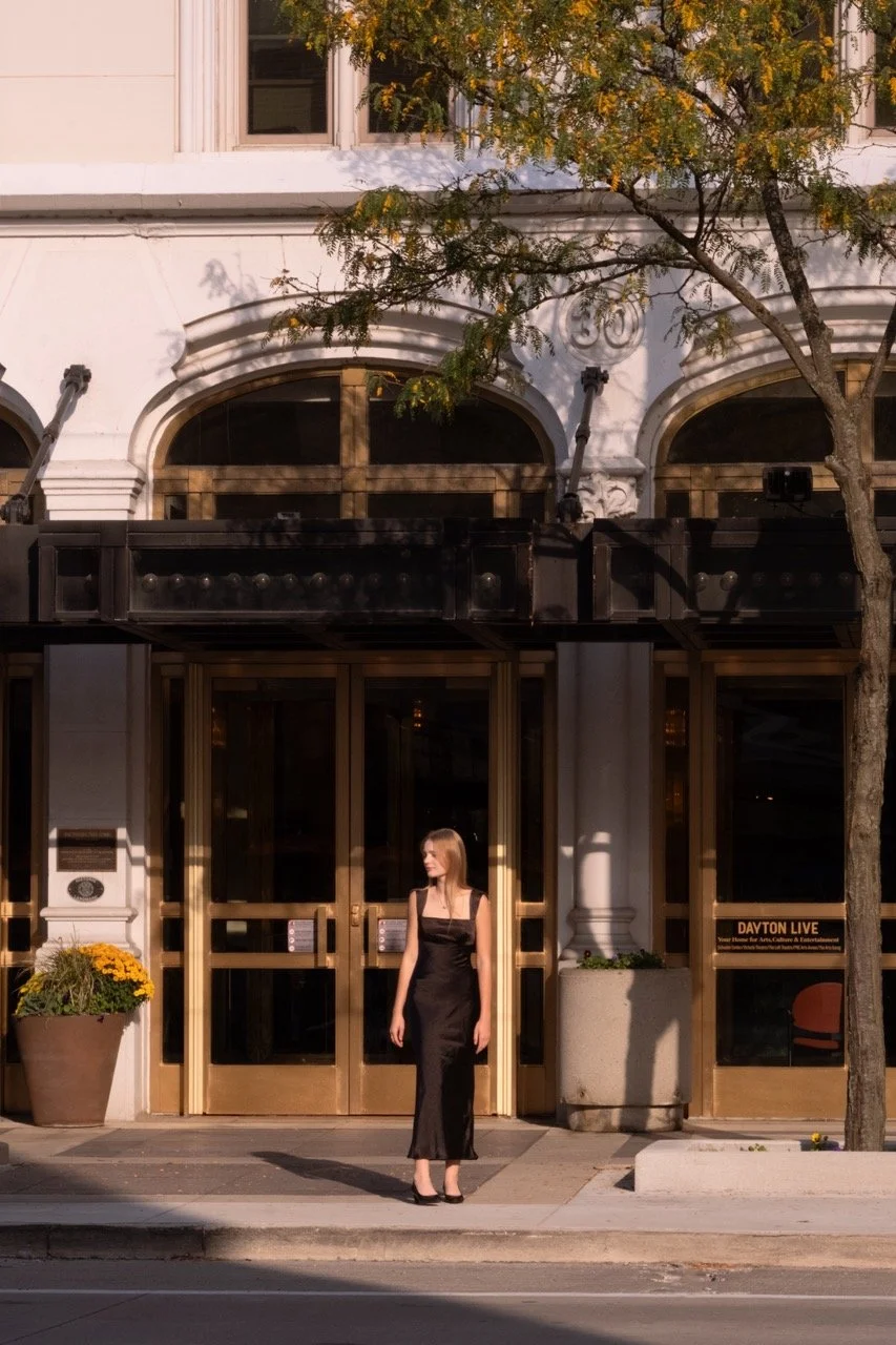A woman in a black dress standing on a city sidewalk in front of a building with large glass doors, a tree with green leaves, and a planters with yellow flowers.