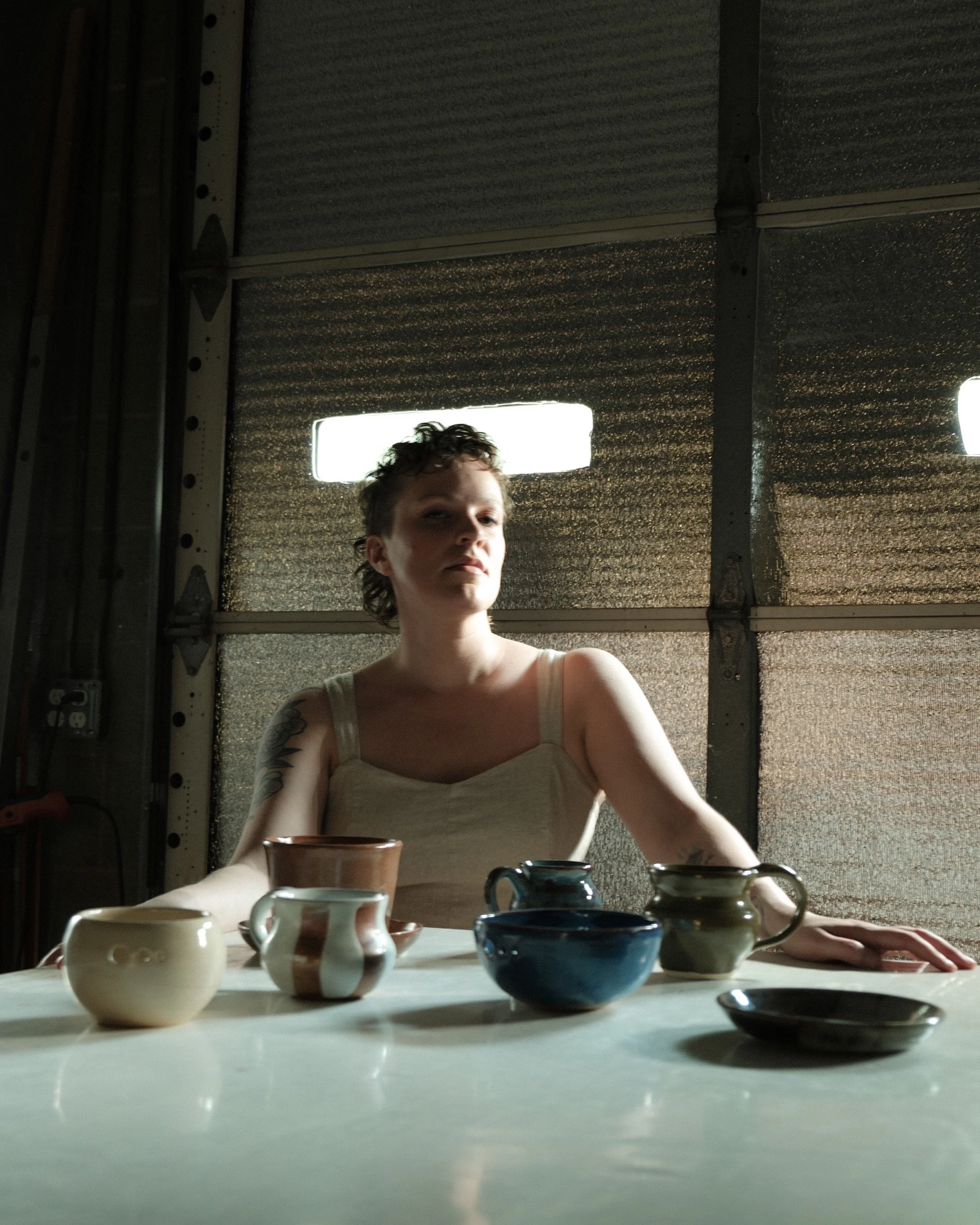 Woman with short curly hair and tattoos on her arms looking down at a table filled with various ceramic cups and bowls in front of her. The background features a textured metal ceiling with rectangular light panels.