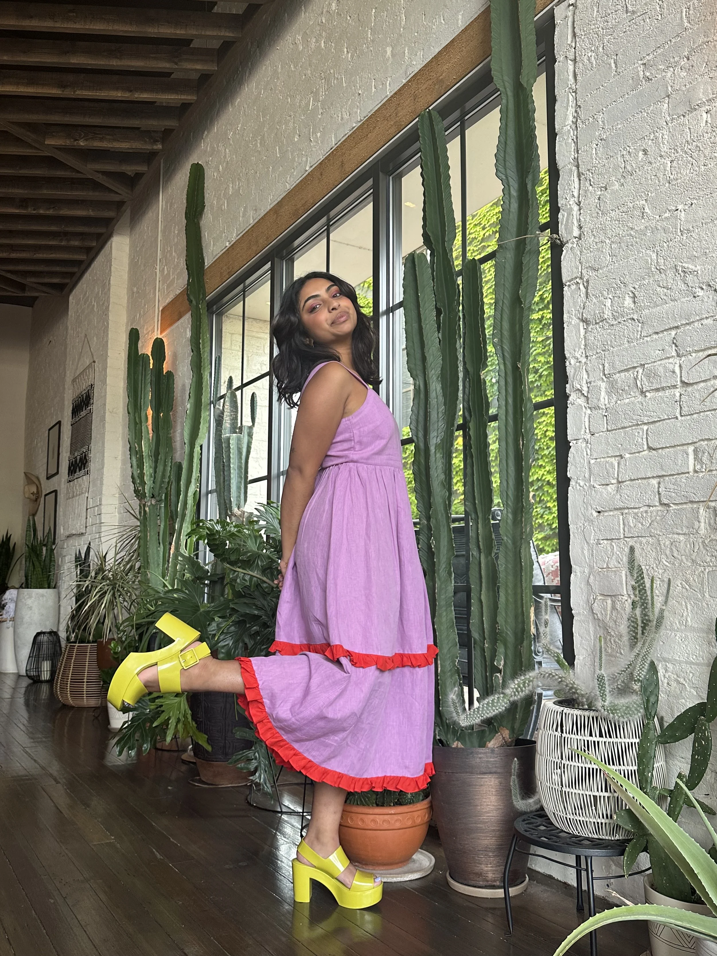 A woman with dark wavy hair wearing a purple dress with red ruffle trim and bright yellow chunky platform shoes standing among various potted green plants indoors, near large windows with black frames and a white brick wall.