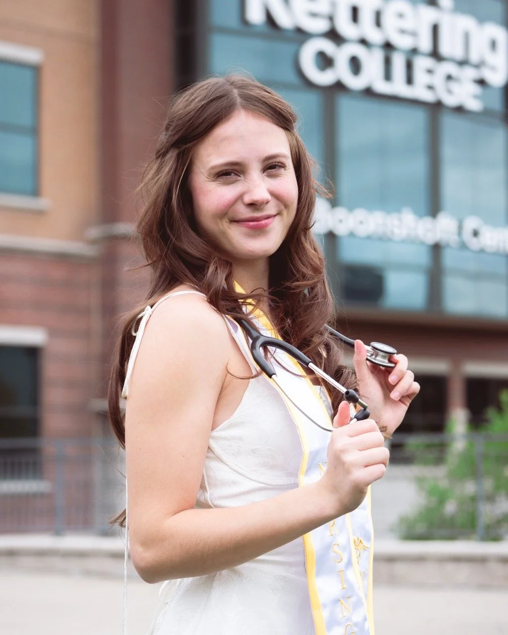 Young woman with long brown hair, wearing a white dress and a graduation stole, holding a stethoscope, smiling in front of a building with a sign that reads 'Rettering College'.