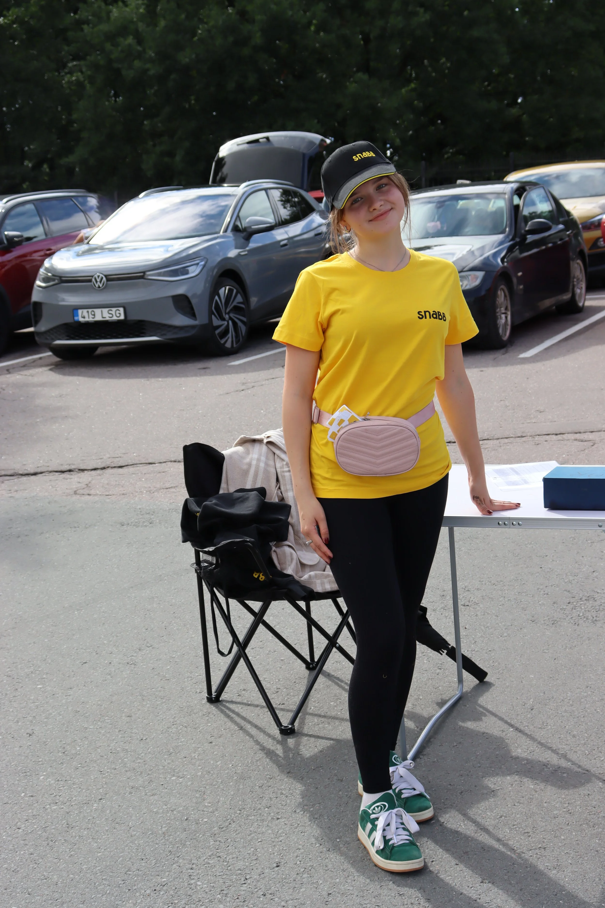 A young woman in a yellow t-shirt and black leggings standing outdoors next to a table and chair. She is wearing a black cap and smiling at the camera. Behind her is a parking lot with several cars and trees in the background.