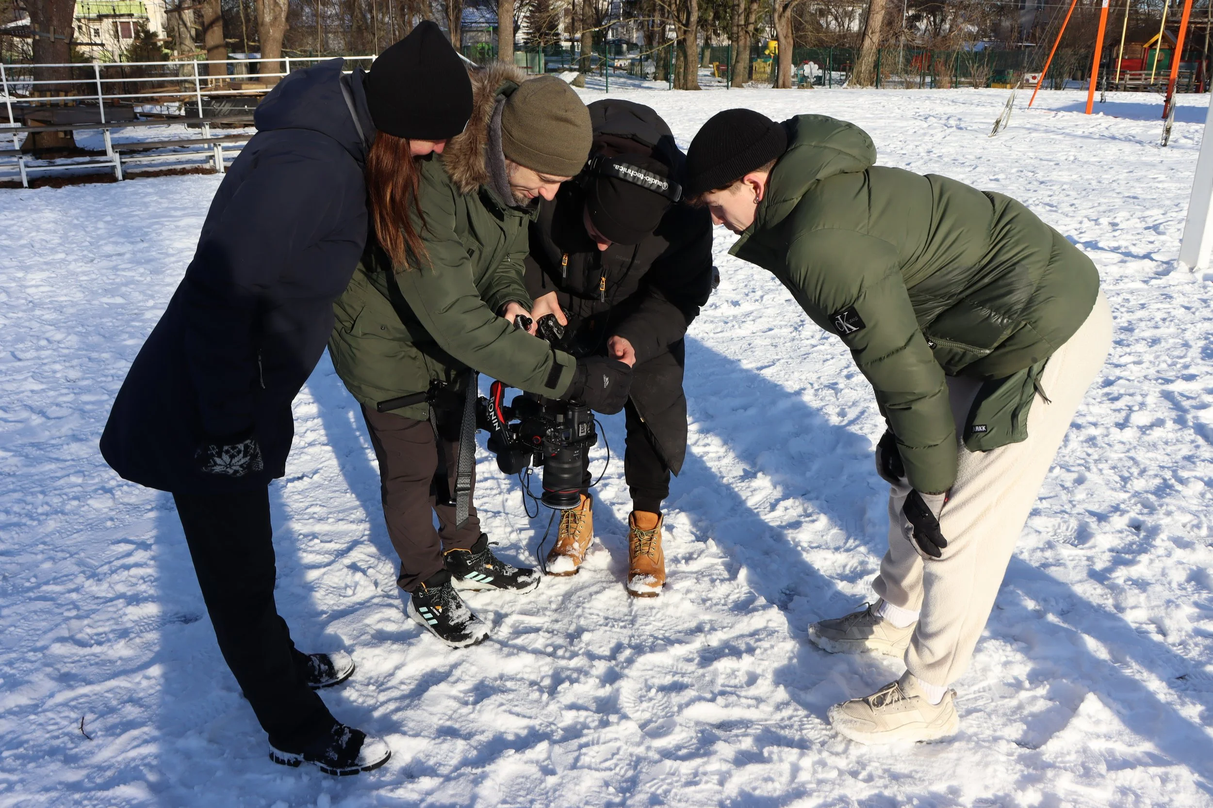 Four people in winter clothing and ice cleats gathered around a camera on snowy ground, looking at something on the camera in an outdoor park with trees and playground equipment in the background.
