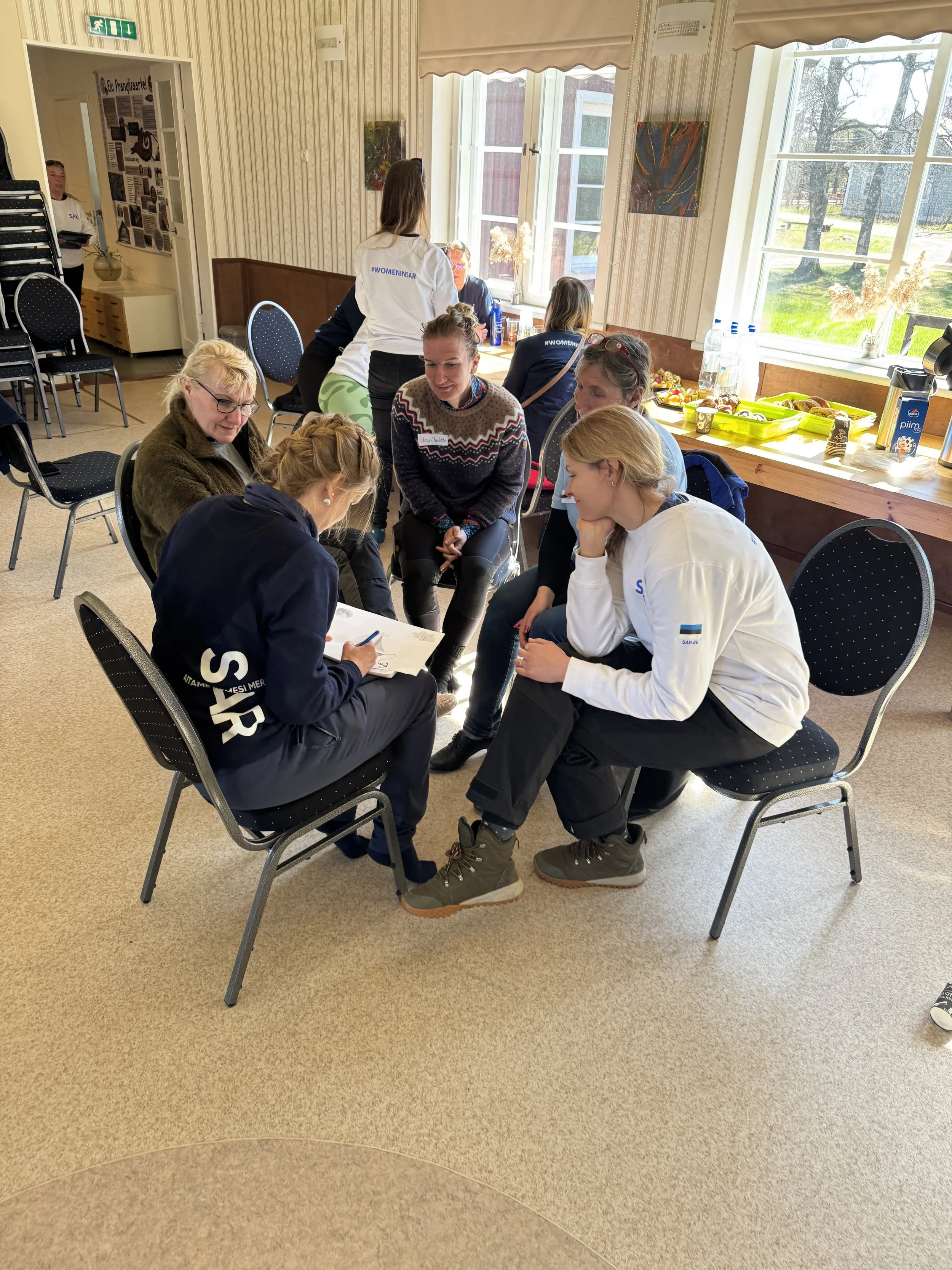 Group of women sitting in a circle in a room with large windows, discussing and taking notes.