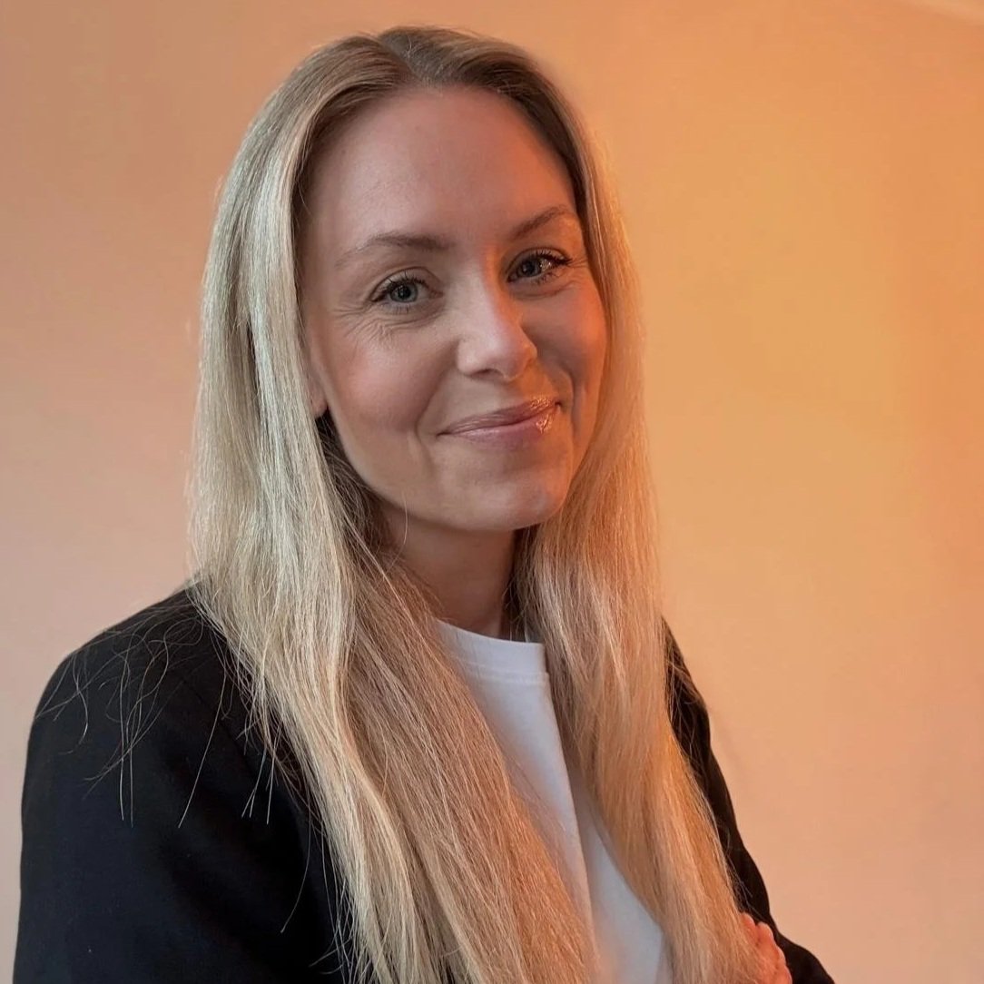 Porträtt av en kvinna med långt blont hår, läppsmil, svart jacka och vit tröja, mot en grå-orange bakgrund.