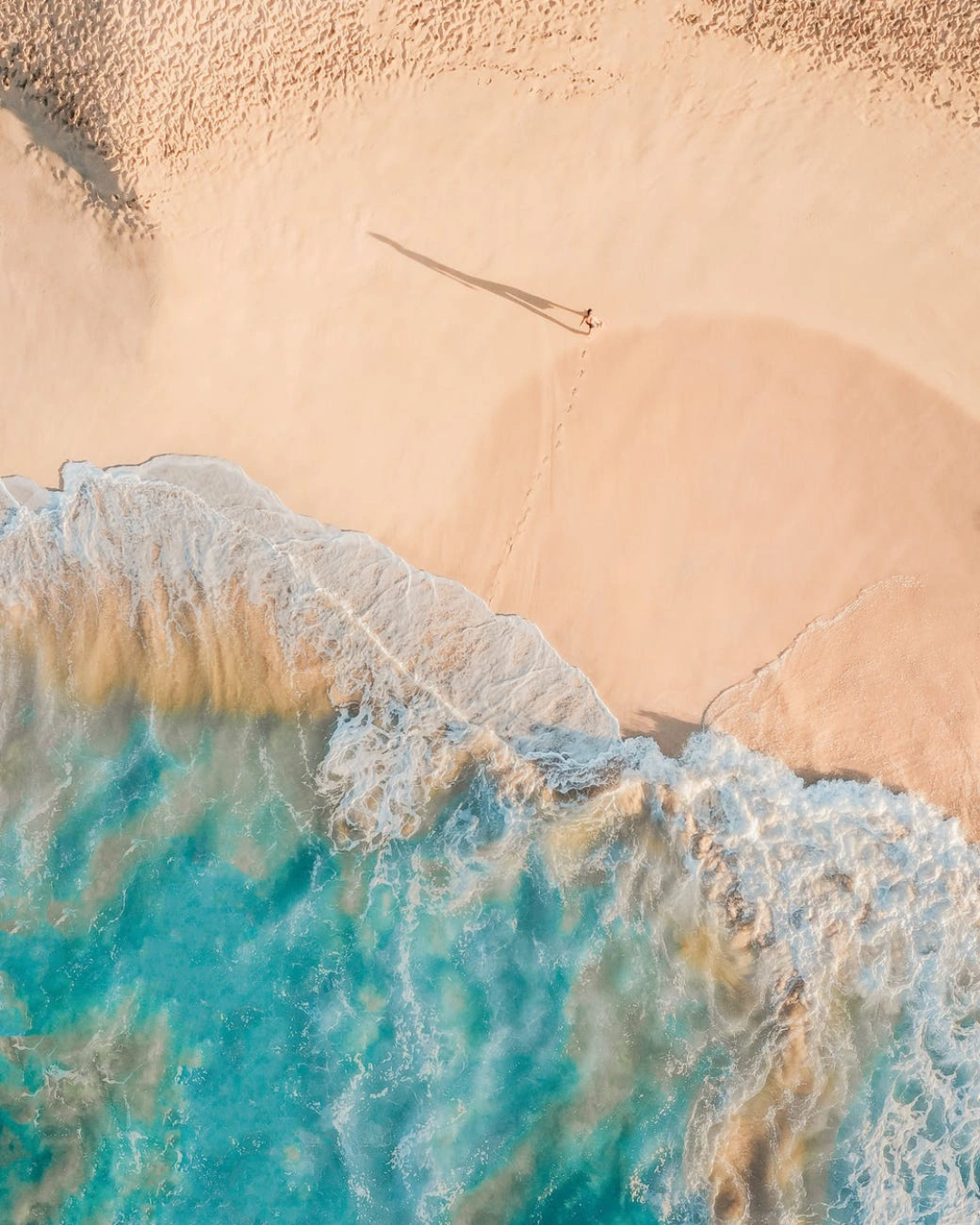 An aerial view of a person lying on a sandy beach near the ocean, casting a long shadow. The beach is mostly empty, with a single set of footprints leading to the person.