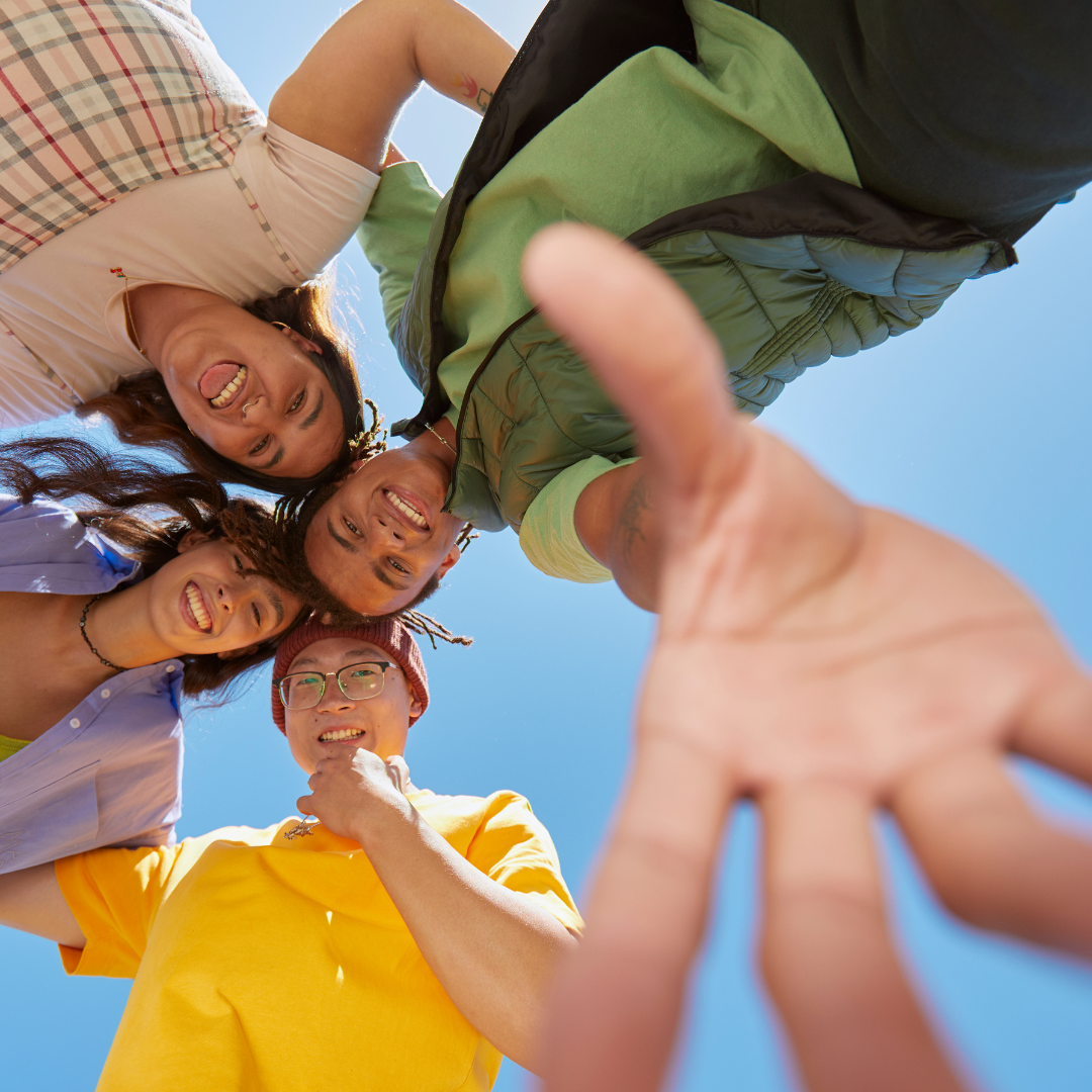 Group of five friends looking down at camera, smiling, with blue sky above, as one person reaches out towards the camera.