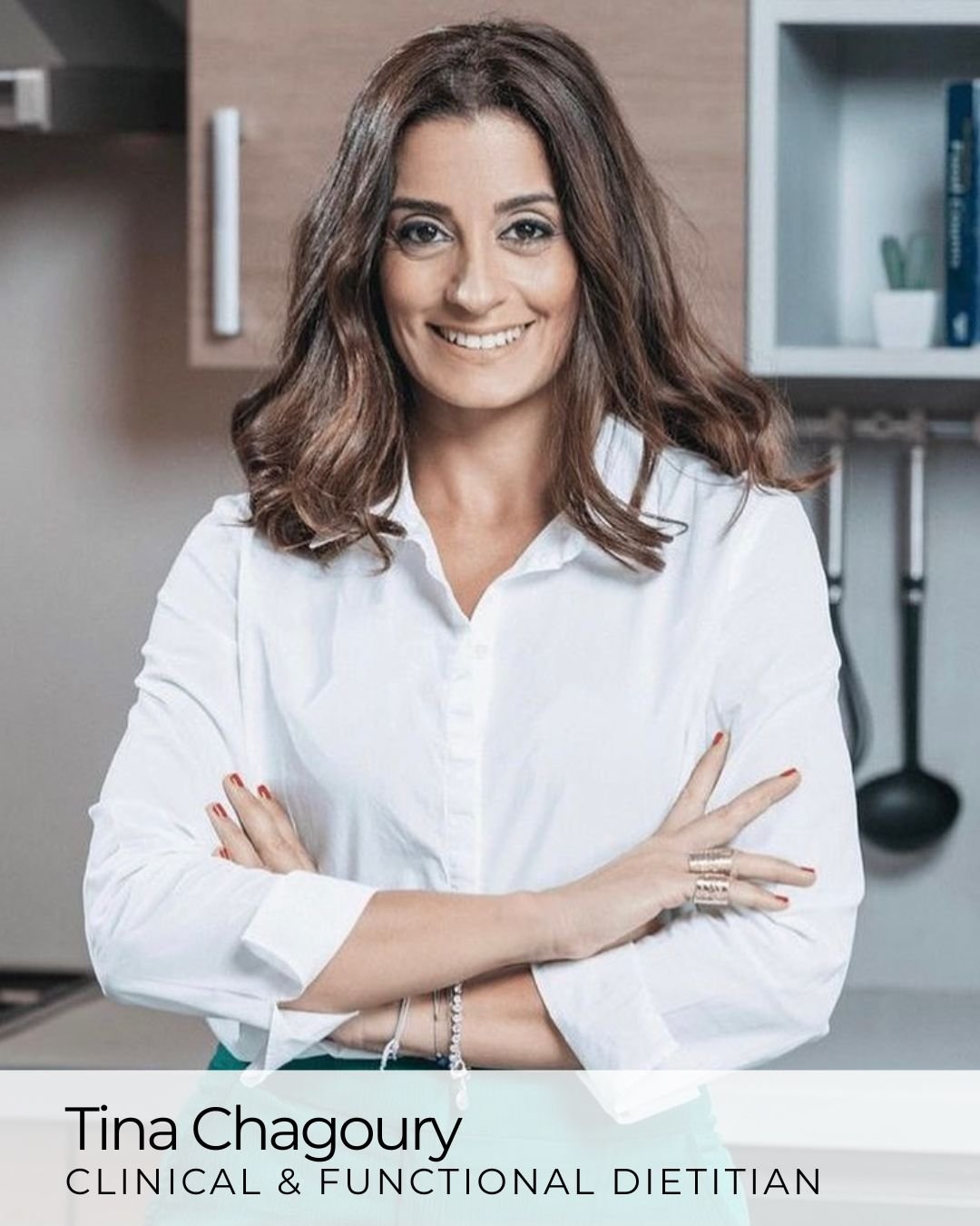 A woman with shoulder-length brown hair smiling with arms crossed, wearing a white shirt and teal pants in a kitchen.