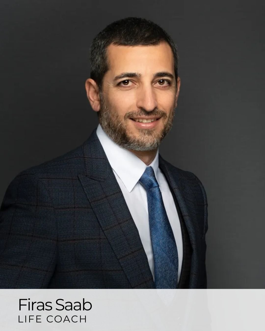 A professional headshot of a man in a dark plaid suit, white shirt, and blue tie, smiling against a dark background.