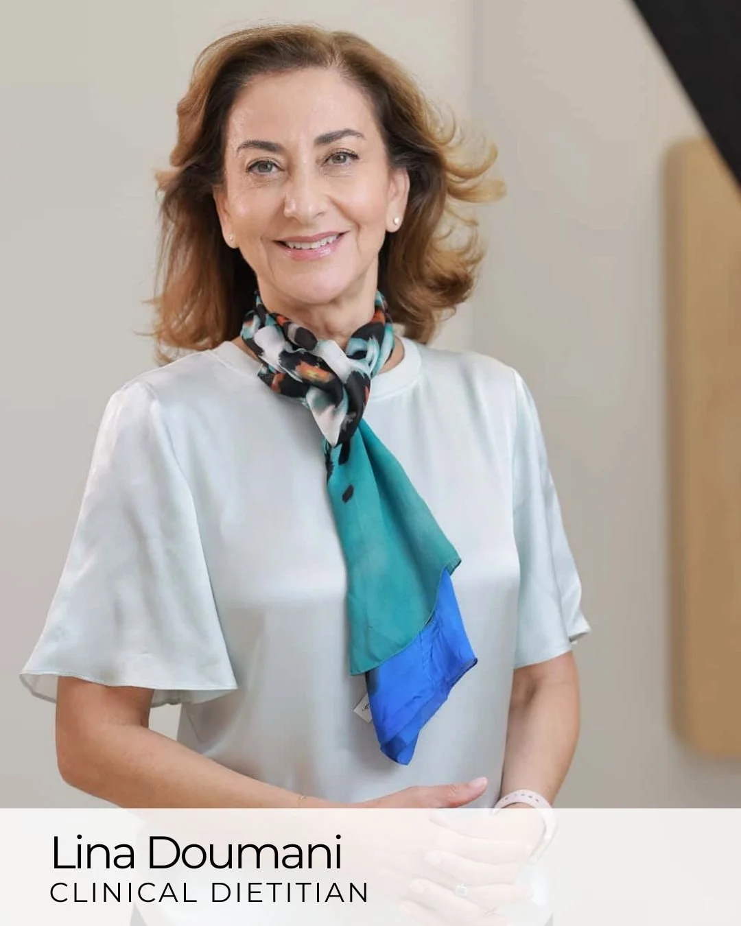 A middle-aged woman with light skin, wavy brown hair, and a smile, standing indoors with a neutral background. She is wearing a white blouse and a colorful silk scarf around her neck.