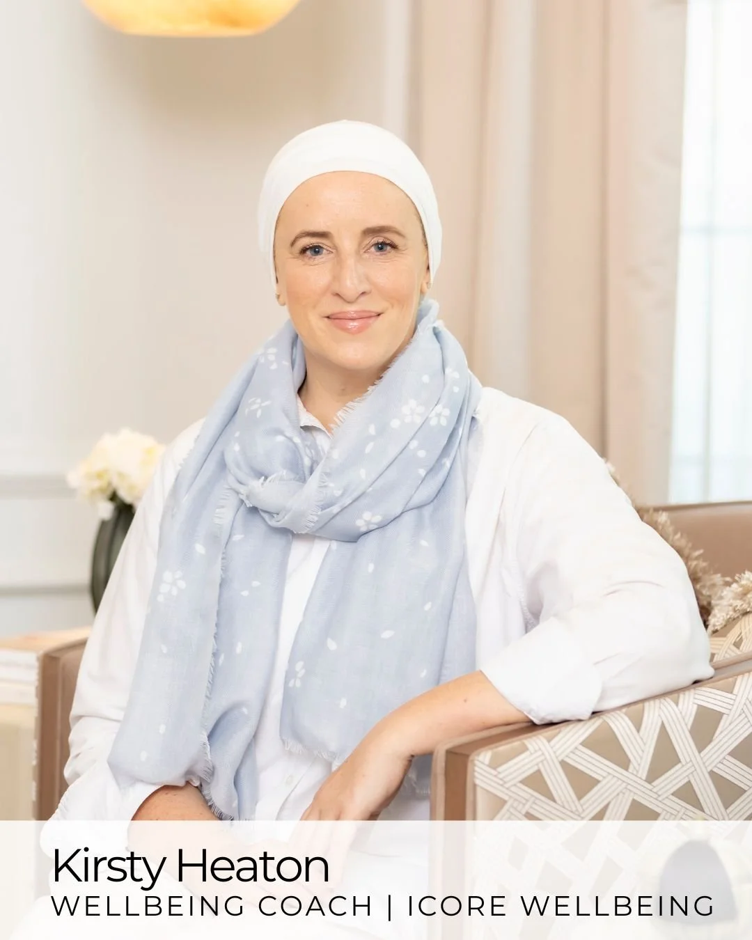 A woman with light skin, blue eyes, and blonde hair, wearing a white headscarf and a light blue scarf, smiling and sitting in a softly lit room with beige curtains and a wooden table in the background.