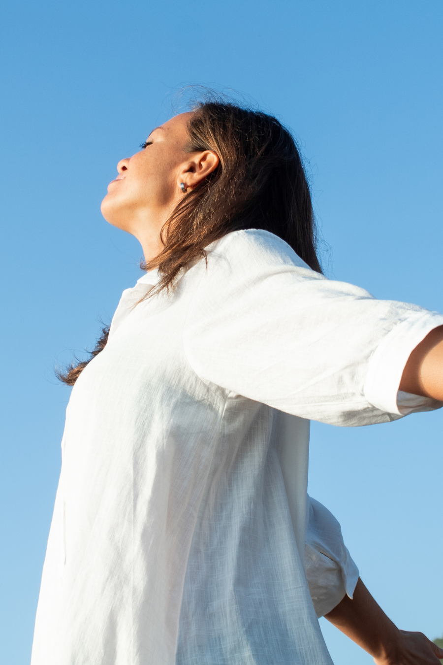 A woman with brown hair in a white shirt standing outdoors with her eyes closed and face tilted upward against a clear blue sky.