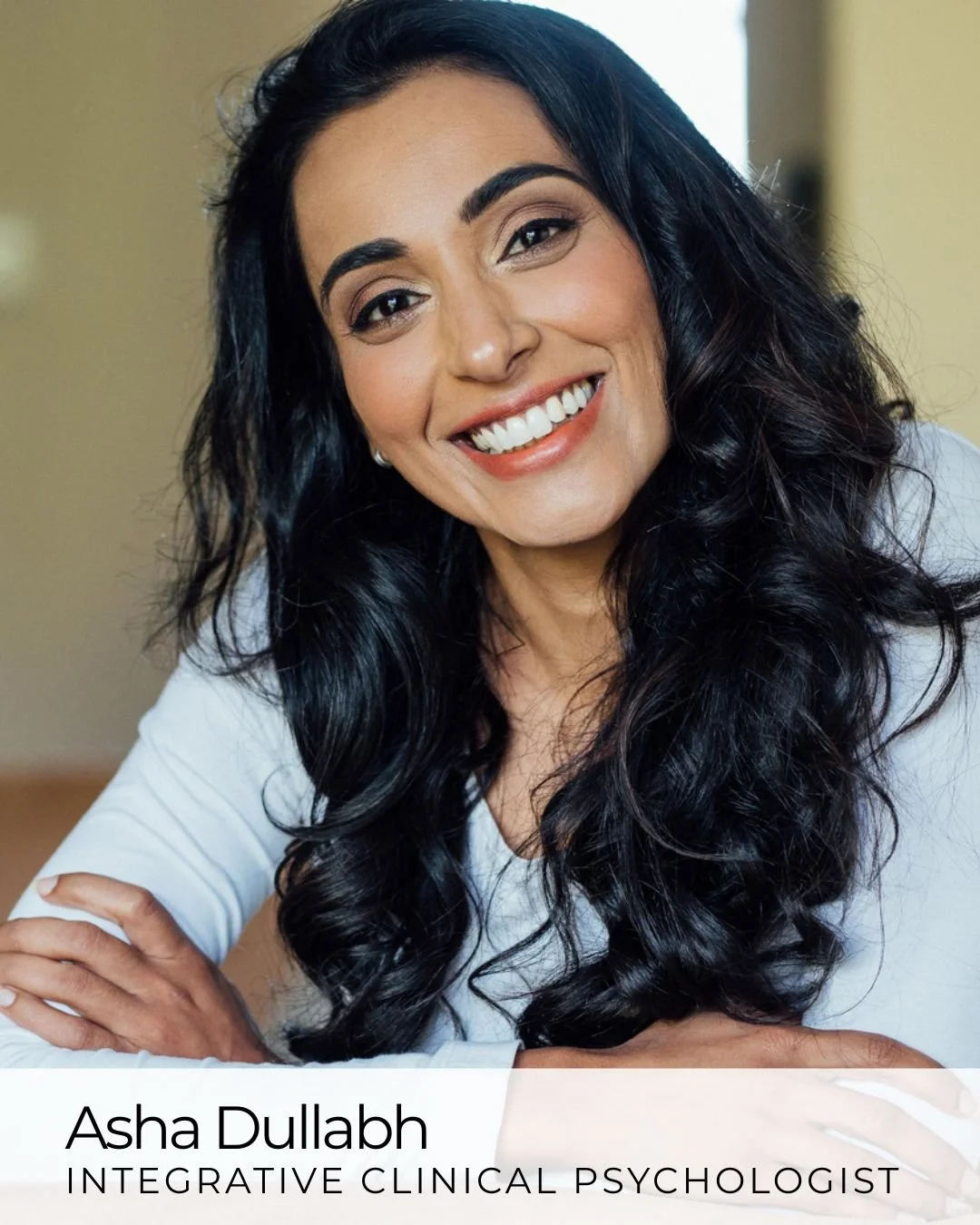A woman with long, curly black hair, wearing a white shirt, smiling brightly with white teeth, sitting at a table with her arms crossed in front of her.